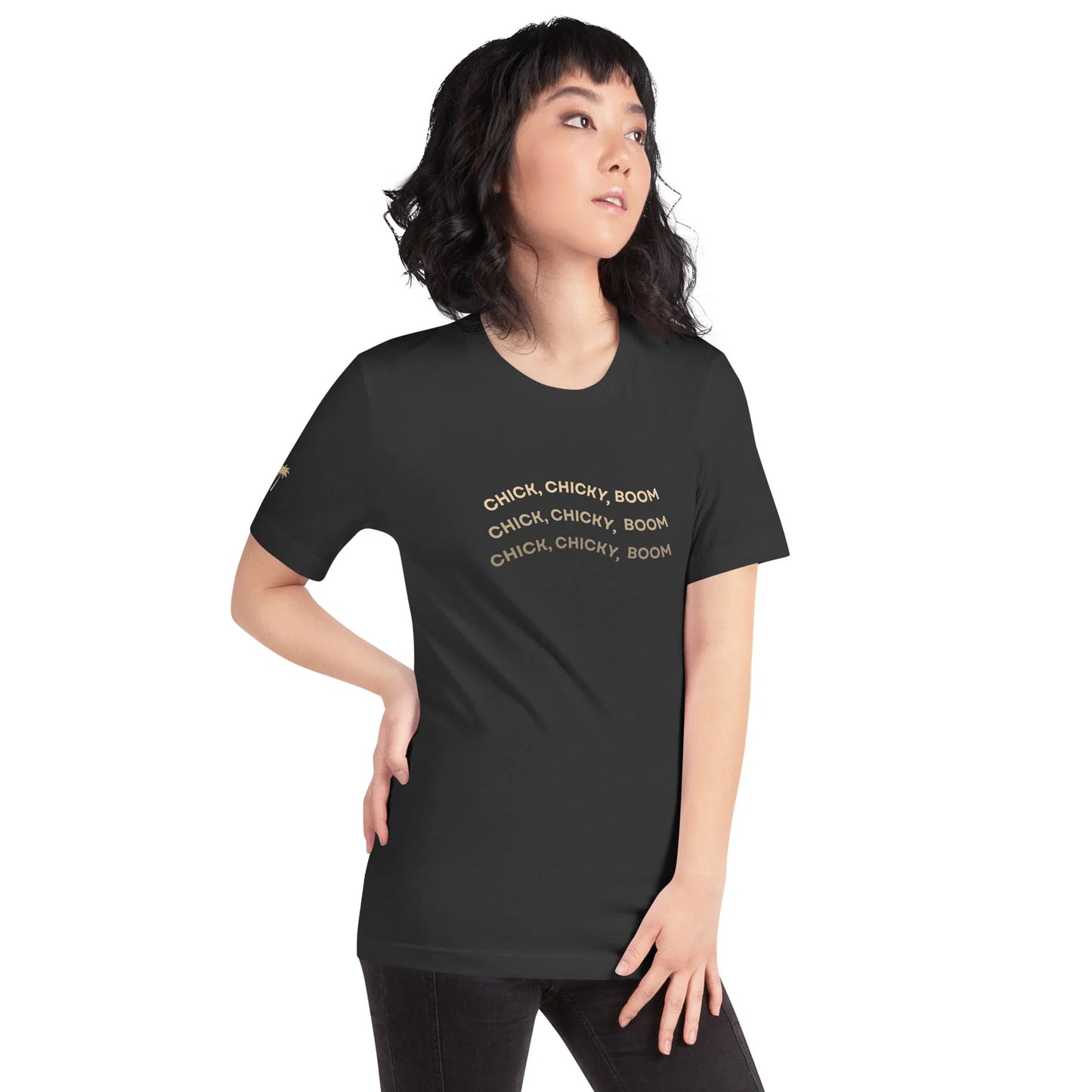 A woman with wavy black hair stands with one hand on her hip, wearing a fitted graphic tee in comfortable cotton that says CHICK, CHICKY, BOOM in gold text, repeated three times. She looks off to the side with a neutral expression.