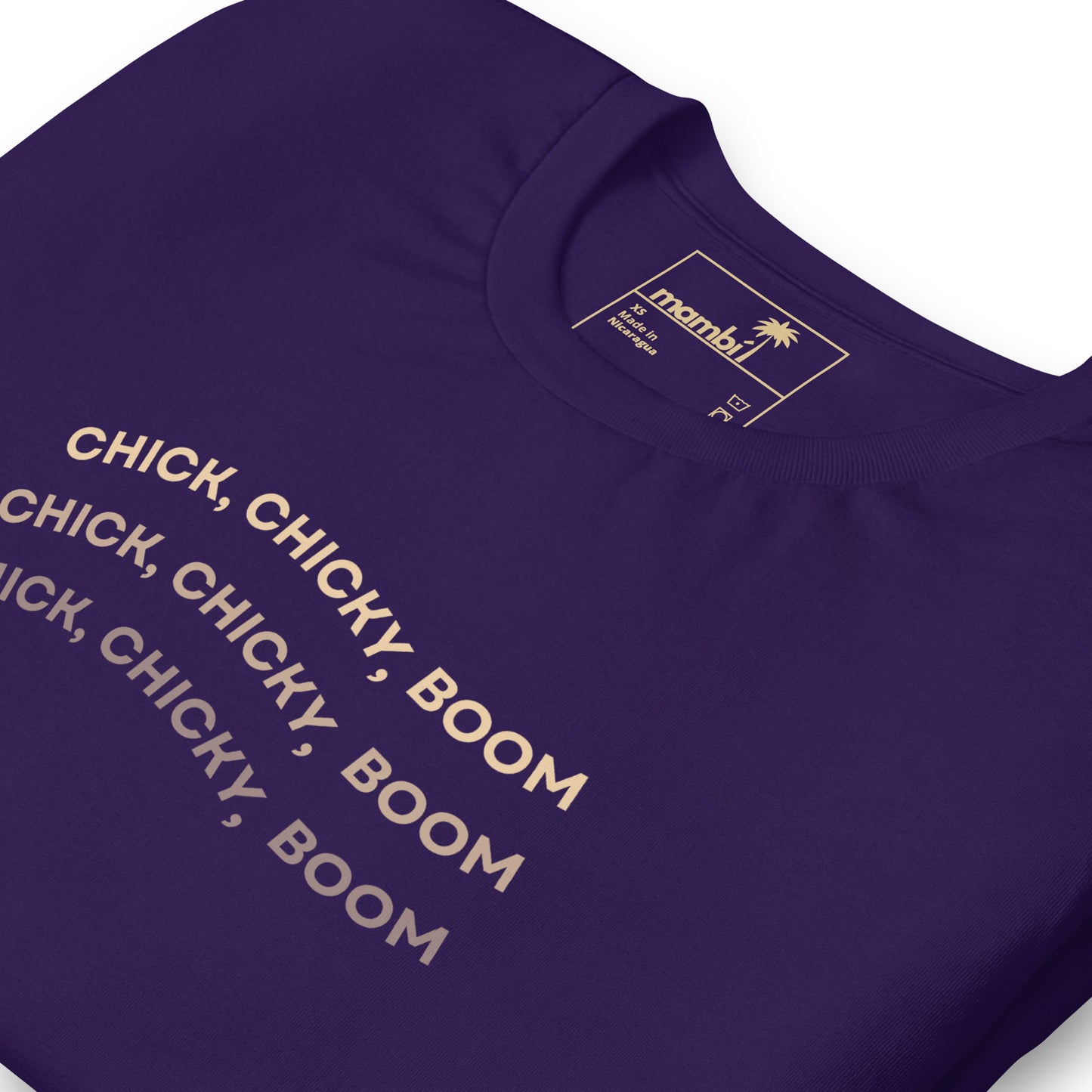 A close-up of a purple graphic tee with the words “CHICK, CHICKY, BOOM” repeated three times in bold, light-colored text. The comfortable cotton shirt’s label at the collar displays the brand name “mambí.”.