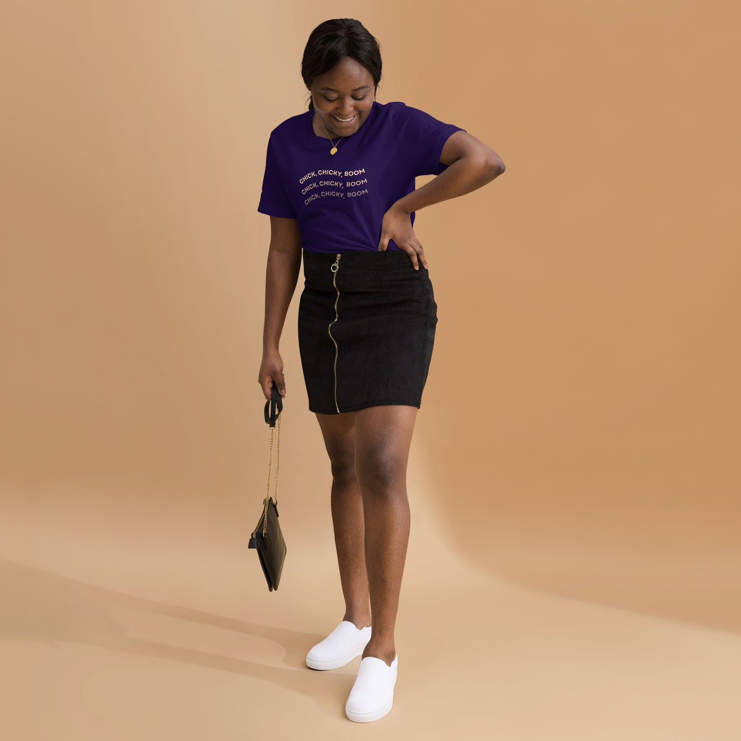 A smiling woman stands against a beige background, wearing a comfortable cotton purple T-shirt, black zip-up skirt, and white slip-on shoes. She holds a small black handbag and looks down at her skirt while touching it.