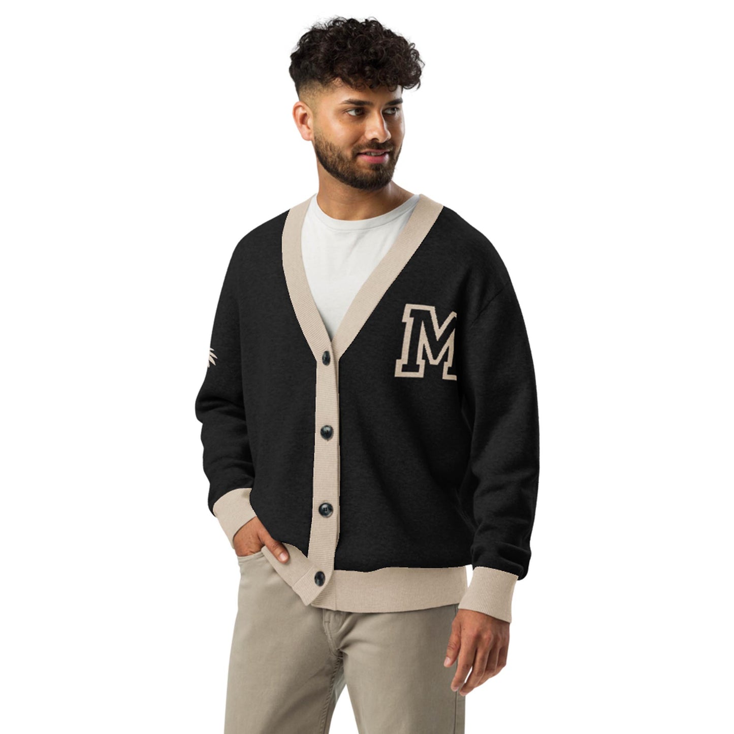A man with curly hair and a beard wears a stylish two-tone cardigan with beige trim and an M patch, paired with light-colored pants. He stands looking to his left with a slight smile.