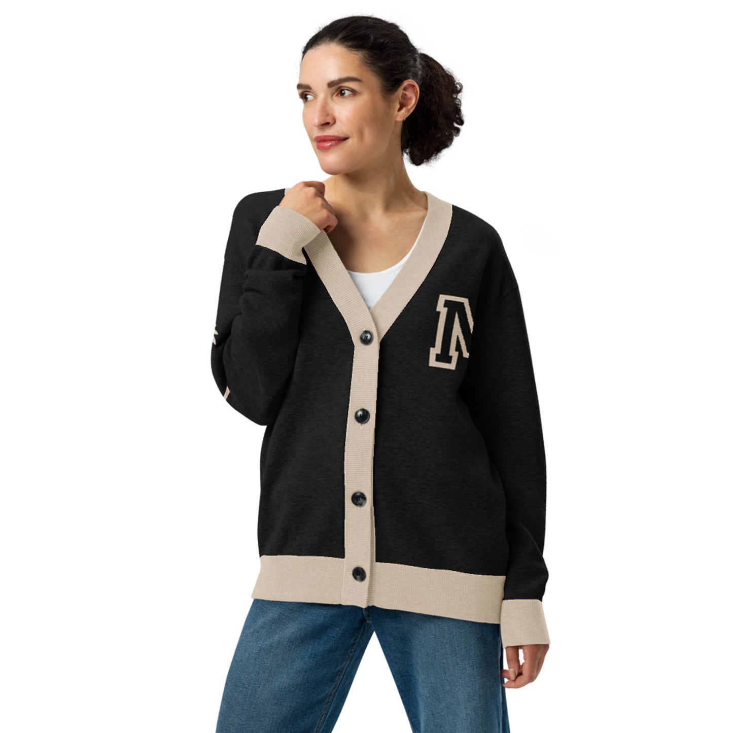 A woman with dark hair in a bun wears a two-tone cardigan with beige trim and buttons, featuring a large A patch on the left side, over a white top and blue jeans. She looks to the side and smiles slightly.
