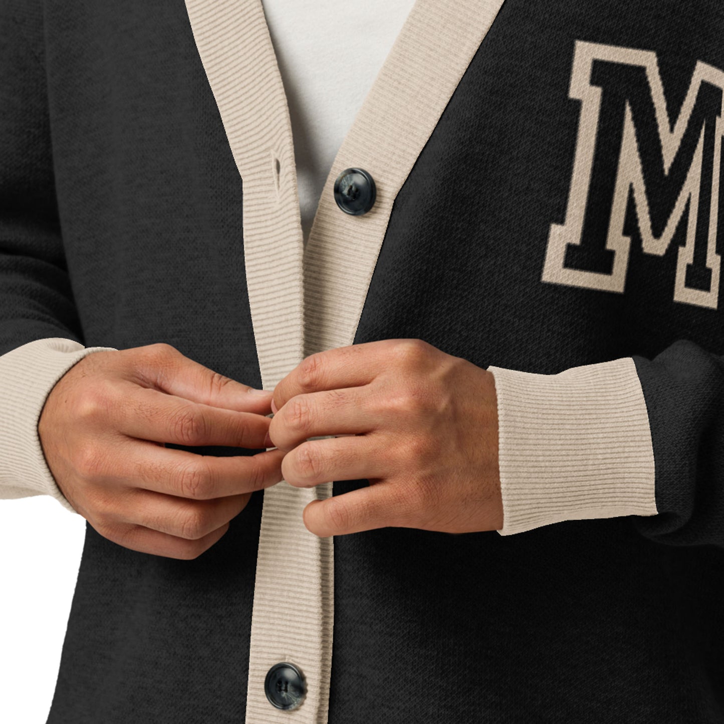 A person buttoning a two-tone cardigan with beige trim and large black buttons, featuring a white letter M patch on the left side. Only their hands and torso are visible.