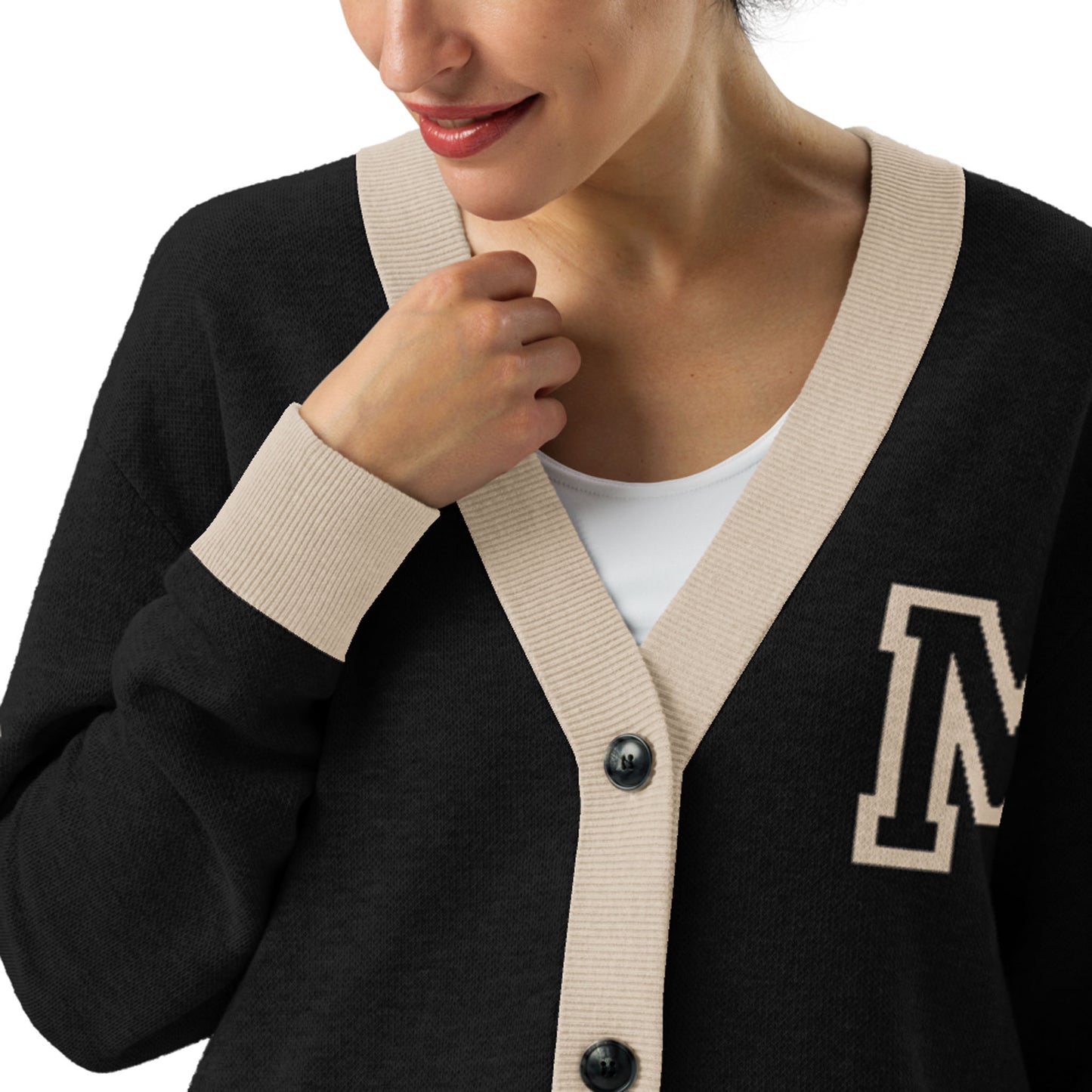 A woman wearing a two-tone cardigan with beige trim and an M patch on the chest, over a white top, lightly touching the collar with her left hand, smiling slightly.
