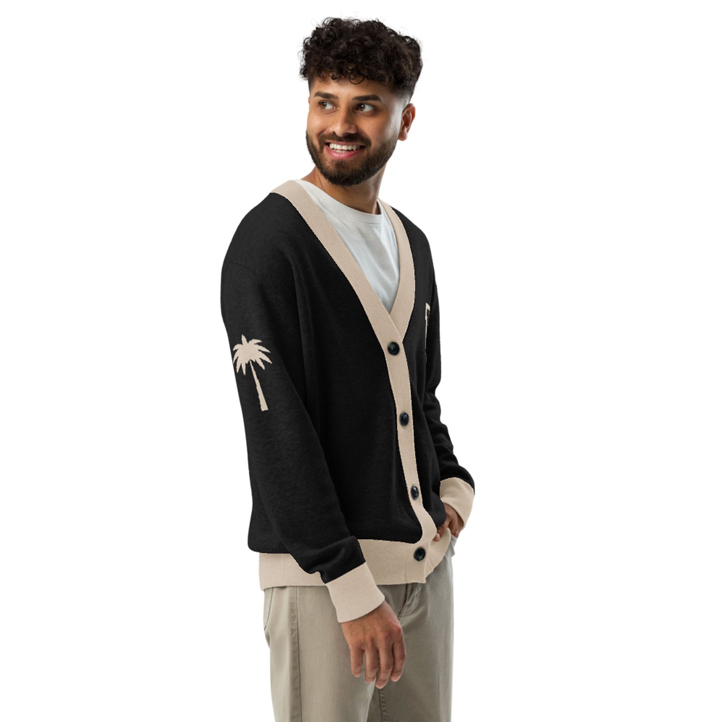 A man with curly hair and a beard smiles while wearing a two-tone cardigan with beige trim and palm tree designs on the sleeve and chest, over a white t-shirt and beige pants. He stands with one hand in his pocket.