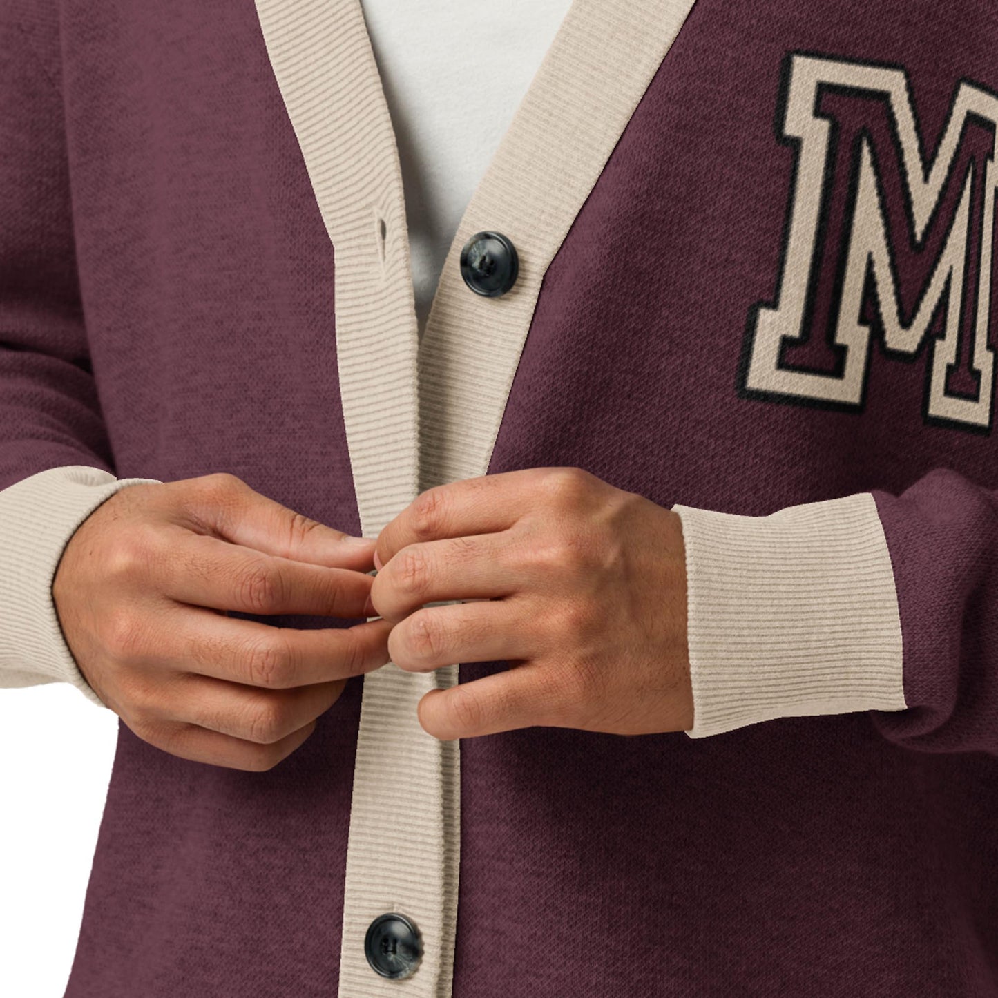 A person buttoning a maroon unisex sweater cardigan with beige trim and black buttons. This lightweight knit features a large, white-outlined letter M patch on the left side, paired with a white shirt underneath.