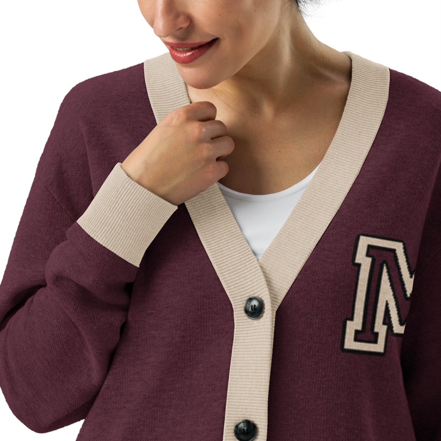 A woman wearing a maroon lightweight knit cardigan with beige trim and an M patch on the left side, over a white top. She is smiling softly and holding the cardigan near the collar.