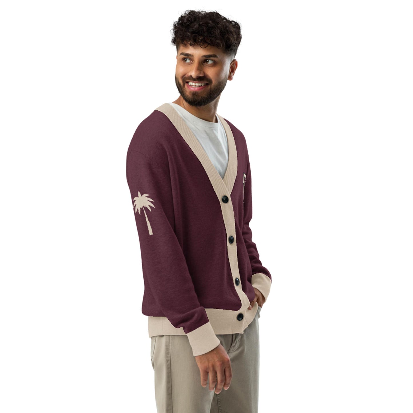 A man with curly hair and a beard smiles while wearing a maroon lightweight knit unisex sweater with beige trim and a palm tree design on the sleeve, a white T-shirt, and beige pants against a white background.