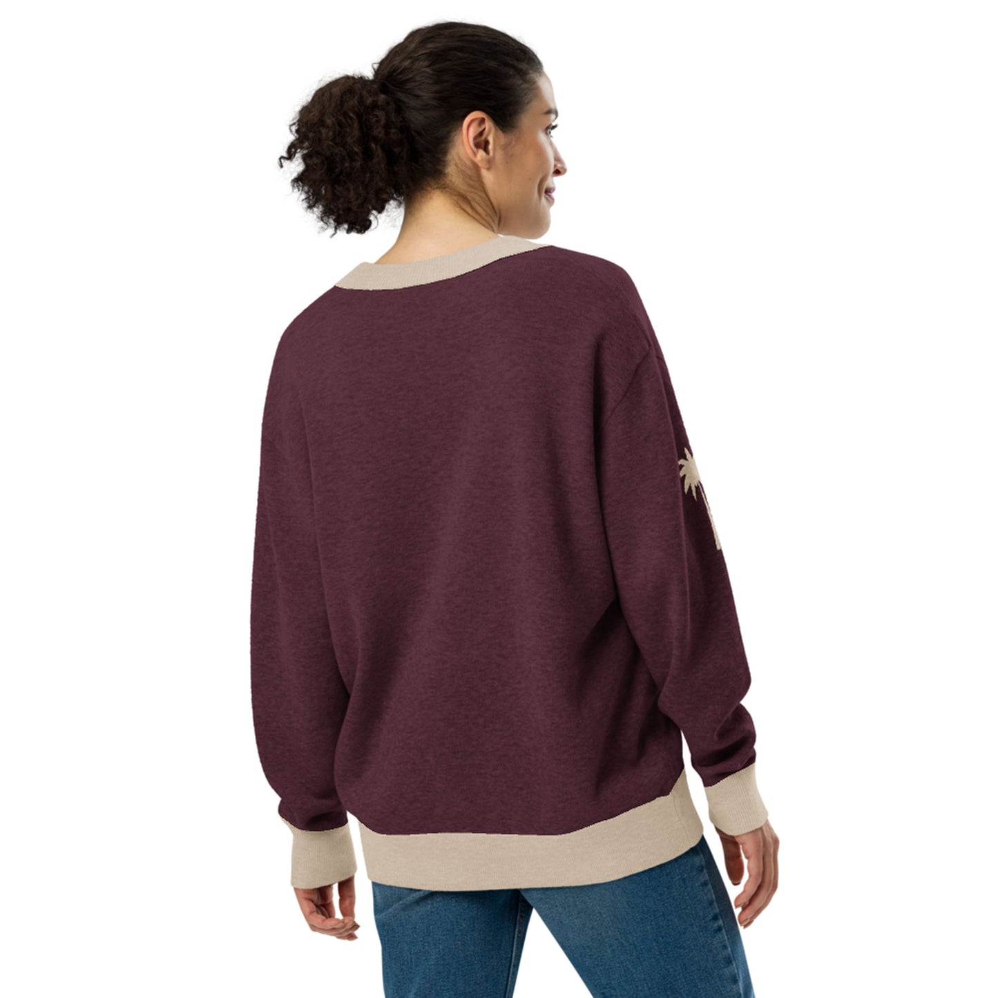 A woman with curly hair in a ponytail wears a loose, maroon two-tone cardigan with beige cuffs and hem, paired with blue jeans. She faces away and smiles, standing against a plain white background.