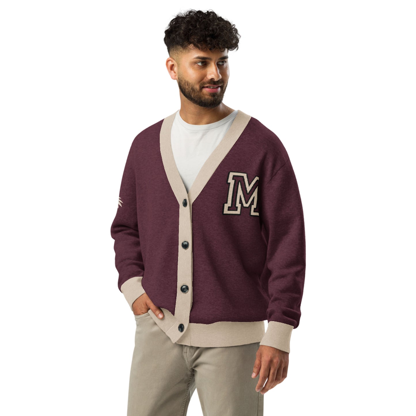 A man with curly hair and a beard wears a two-tone cardigan with beige trim and an M patch over a white t-shirt and beige pants, standing with one hand in his pocket.