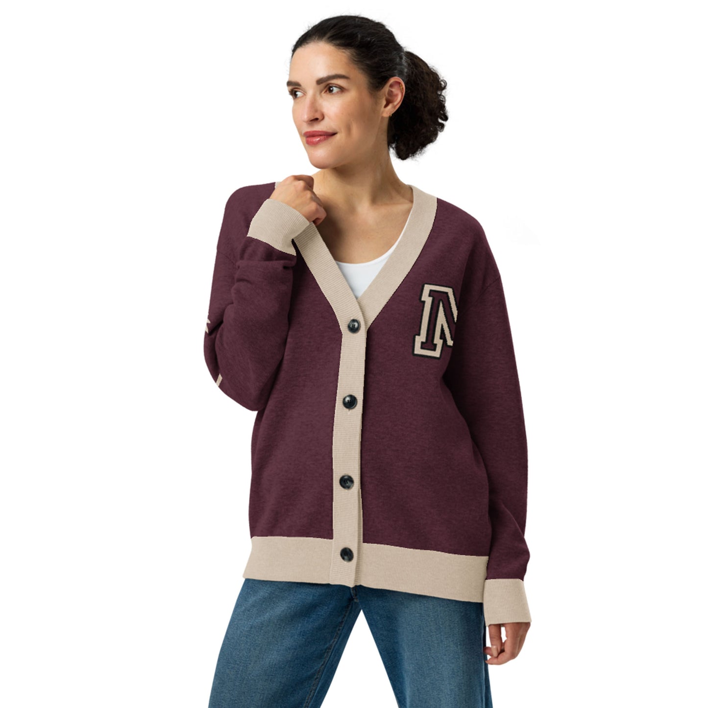 A woman wears a two-tone cardigan with beige trim and black buttons, featuring a large A patch on the left chest. Her hair is tied back as she poses, one hand touching the collar and a slight smile on her face.