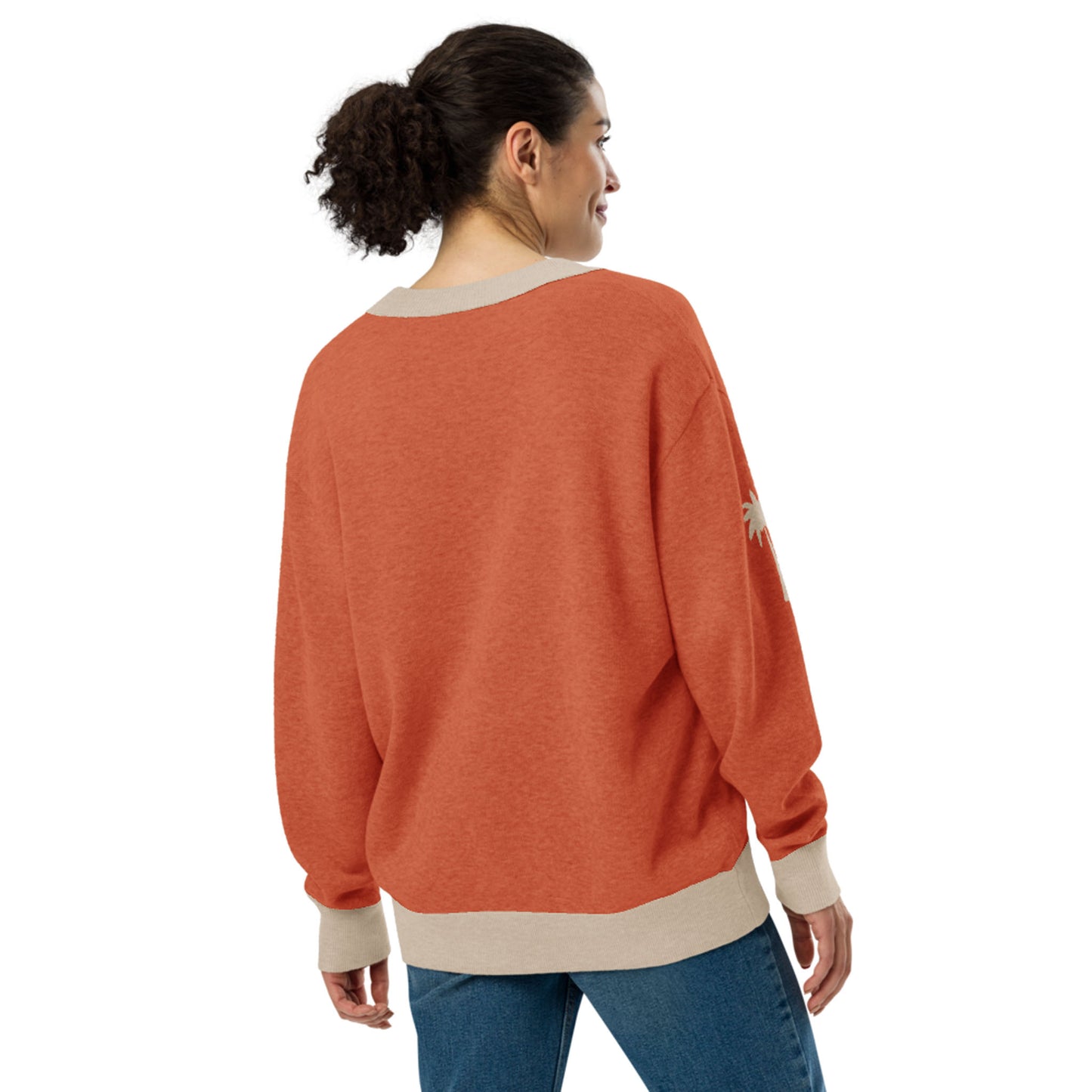 A woman with curly hair in a ponytail is wearing an orange and beige two-tone cardigan with blue jeans, standing and smiling while looking to her left, shown from the back against a white background.