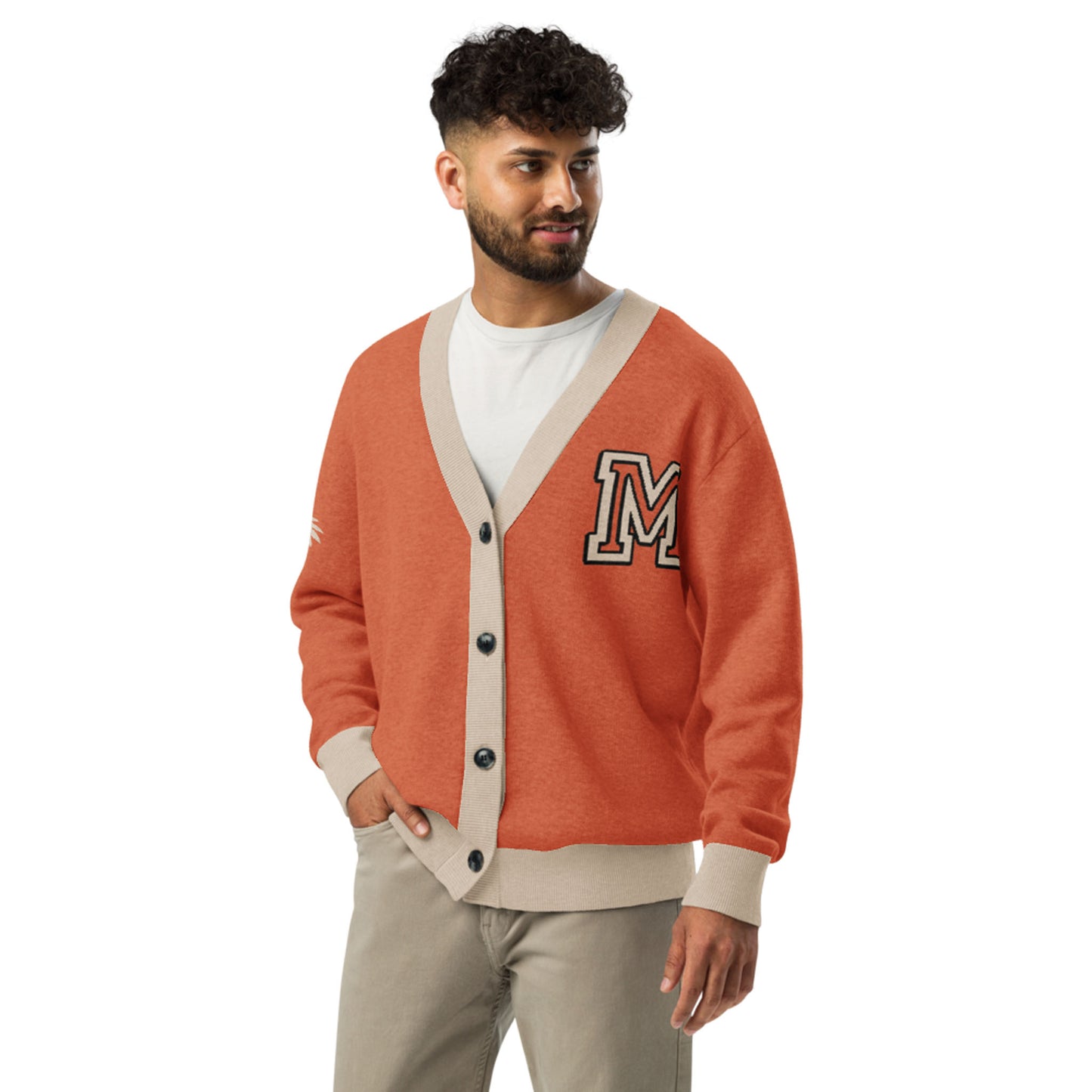 A man with curly hair and a beard wears a stylish two-tone cardigan in burnt orange with beige trim and a large letter M patch. One hand is in his pocket as he looks to the side, smiling. This unisex cardigan adds flair to any outfit.