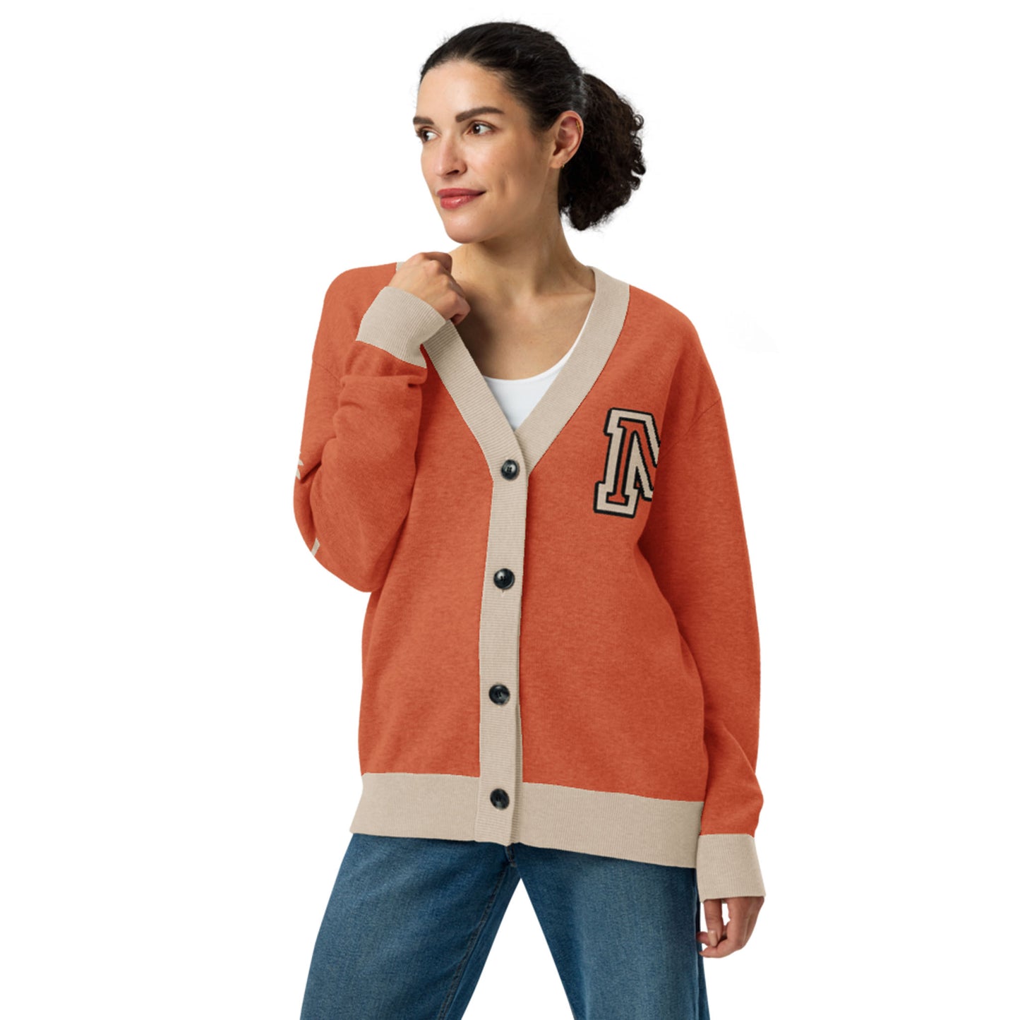 A woman in a two-tone cardigan with rust-orange and beige trim, black buttons, and a letter “A” patch stands looking to the side. She has dark hair tied back, pairing the cardigan with blue jeans and a white shirt.