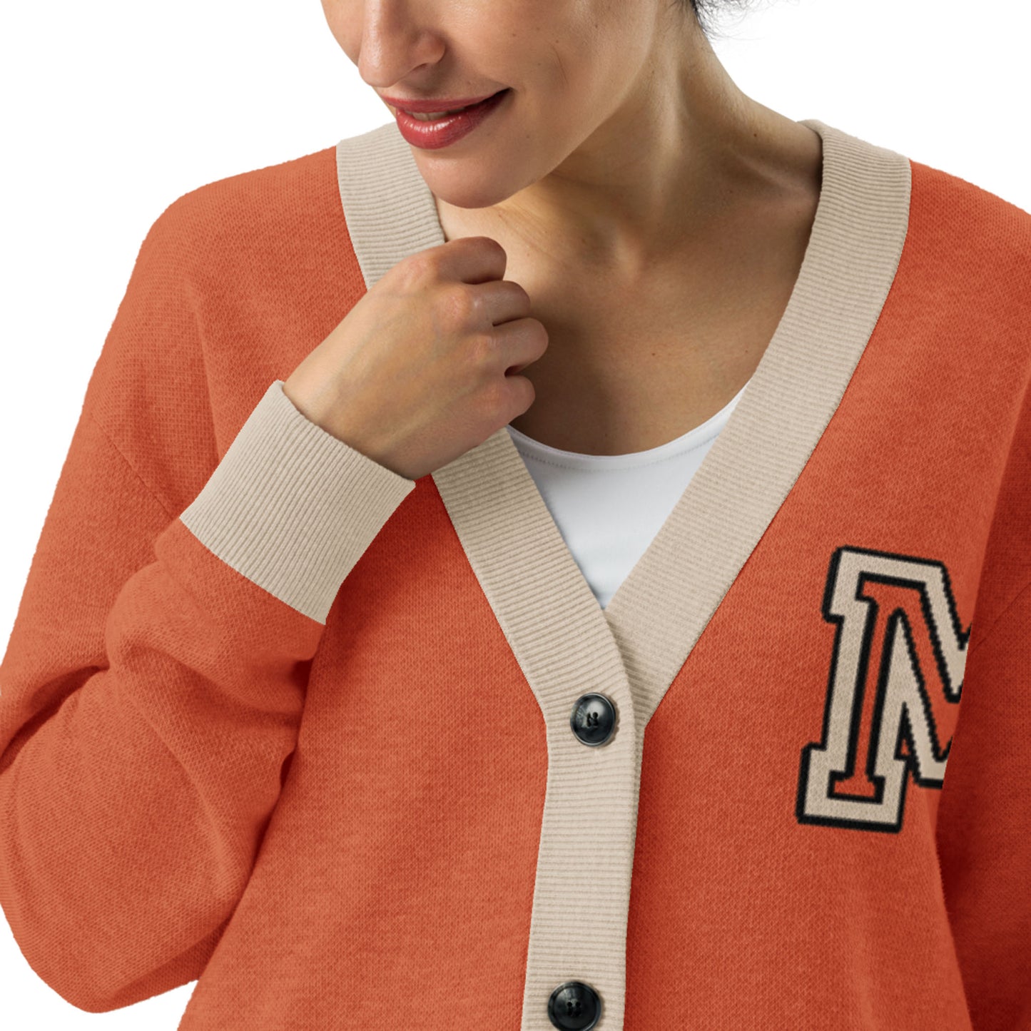 A woman wearing a stylish two-tone cardigan with beige trim and a large letter M patch on the left side, over a white top. She is smiling and holding the cardigan near the collar.
