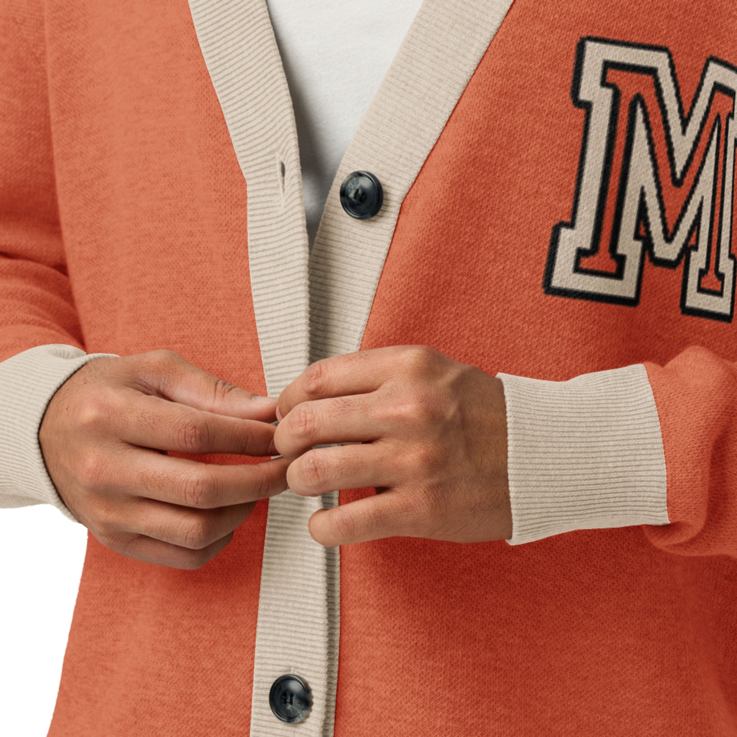 A person wearing a two-tone cardigan—orange with light beige trim and large black buttons—adjusts the front. The unisex cardigan features a bold patch with the letter M on the left side.