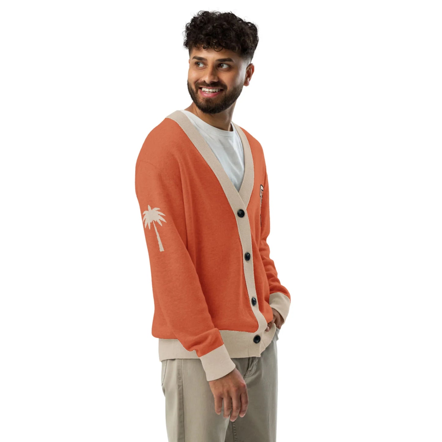 A smiling man wears a white T-shirt, beige pants, and a stylish two-tone cardigan with beige trim and a small white palm tree graphic on the sleeve, standing against a plain white background.