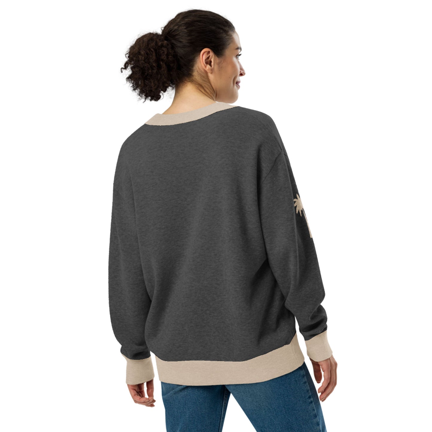 A woman with curly hair in a ponytail is standing and facing away, wearing a loose two-tone cardigan with beige cuffs and hem, paired with blue jeans.