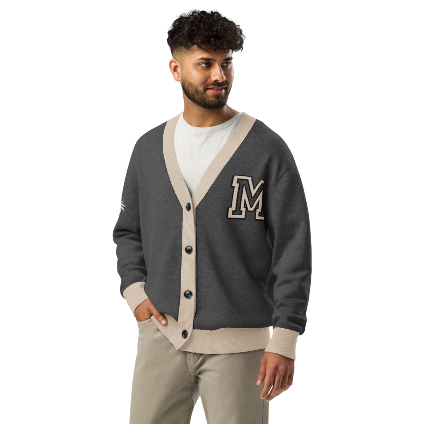 A man with short curly hair and a beard wears a two-tone cardigan with beige trim and a large letter M on the chest. He stands looking to the side, one hand in his pocket, showcasing this stylish unisex cardigan.