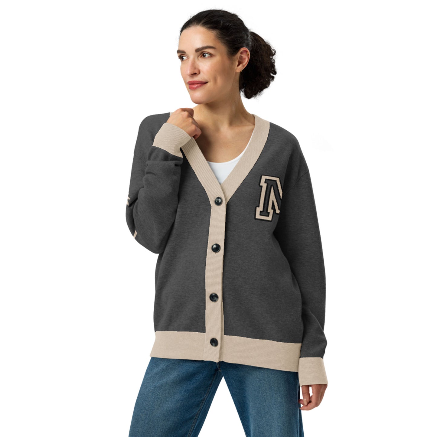 A woman with curly hair in a ponytail wears a two-tone cardigan with beige trim and an A patch, over a white top and blue jeans, standing and looking to the side with a slight smile.