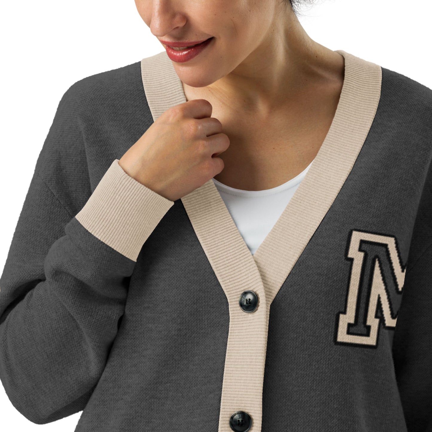 A woman wearing a two-tone cardigan with beige trim and an M patch on the chest over a white top. She is smiling slightly and holding the edge of the cardigan near the collar.