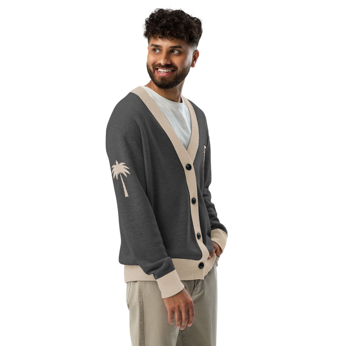 A man with curly hair and a beard smiles while wearing a two-tone unisex cardigan featuring beige trim and a palm tree design on the sleeve, layered over a white shirt and beige pants, standing against a white background.