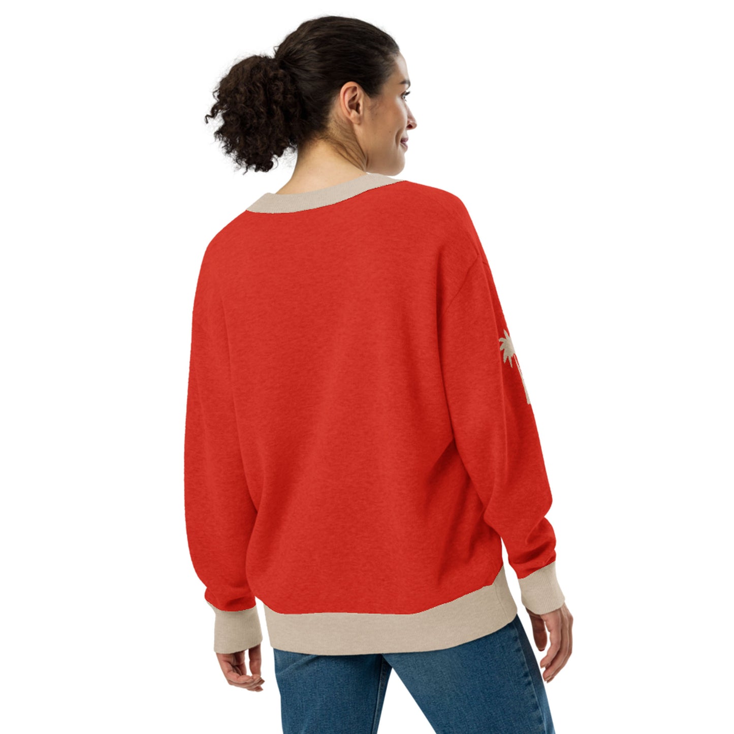 A woman with curly hair in a ponytail is wearing a loose-fitting red two-tone cardigan with beige cuffs and hem, paired with blue jeans. She stands and looks to the side, shown from the back.