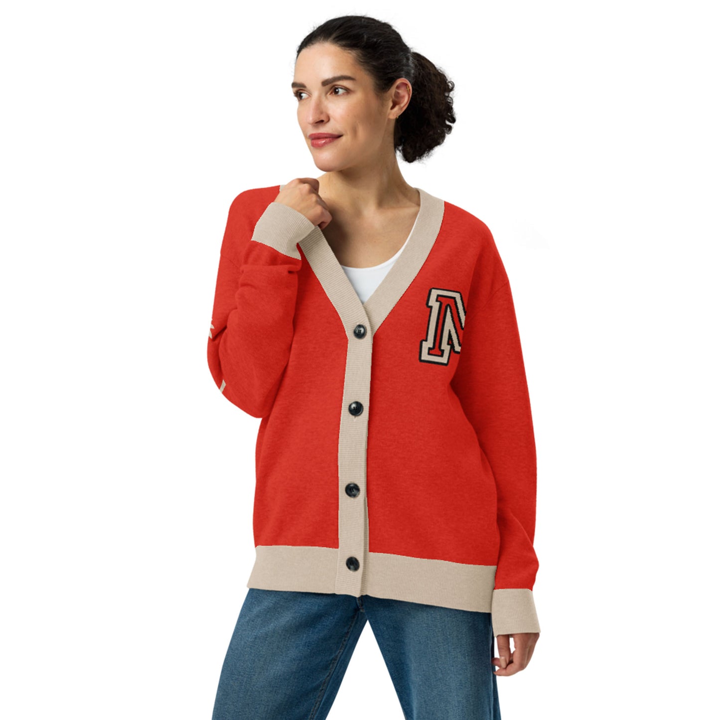 A woman wearing a red two-tone cardigan with beige trim and black buttons, featuring a large letter A patch on the left chest, stands with one hand touching her collar and looks to the side. She has dark hair tied back and wears blue jeans.