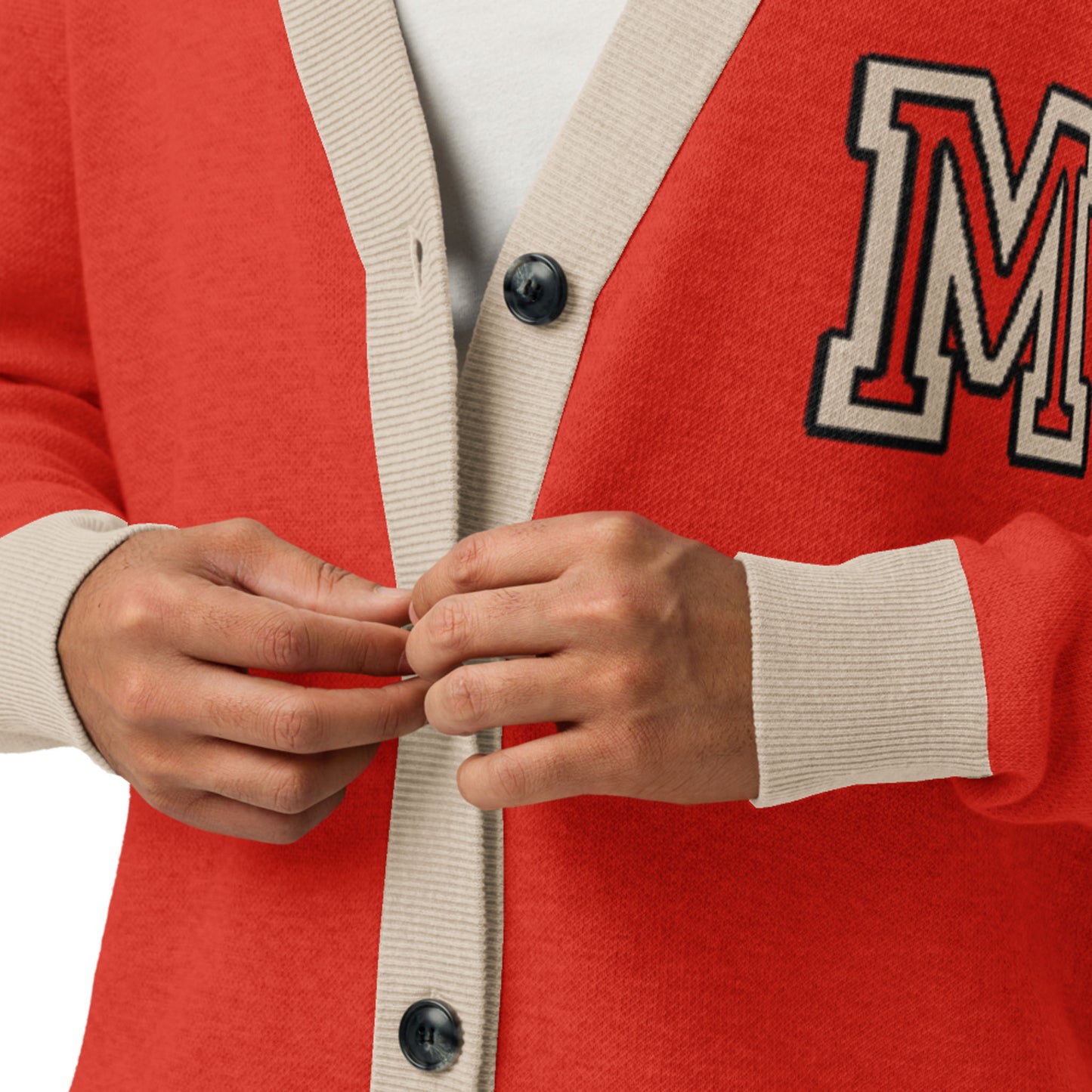 A person wearing a red two-tone cardigan with beige trim and a letter M patch adjusts the front buttons. The unisex cardigan is styled over a white shirt underneath.