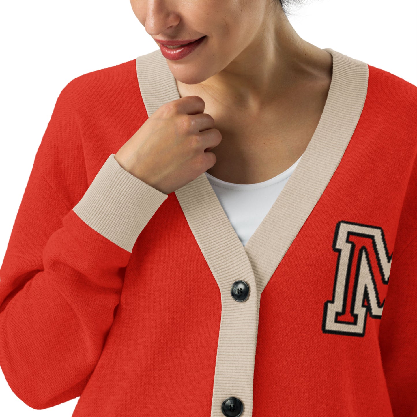 A woman wears a two-tone cardigan with beige trim and an M patch on the chest. She smiles softly, holding the collar of the cardigan, revealing a white top underneath.