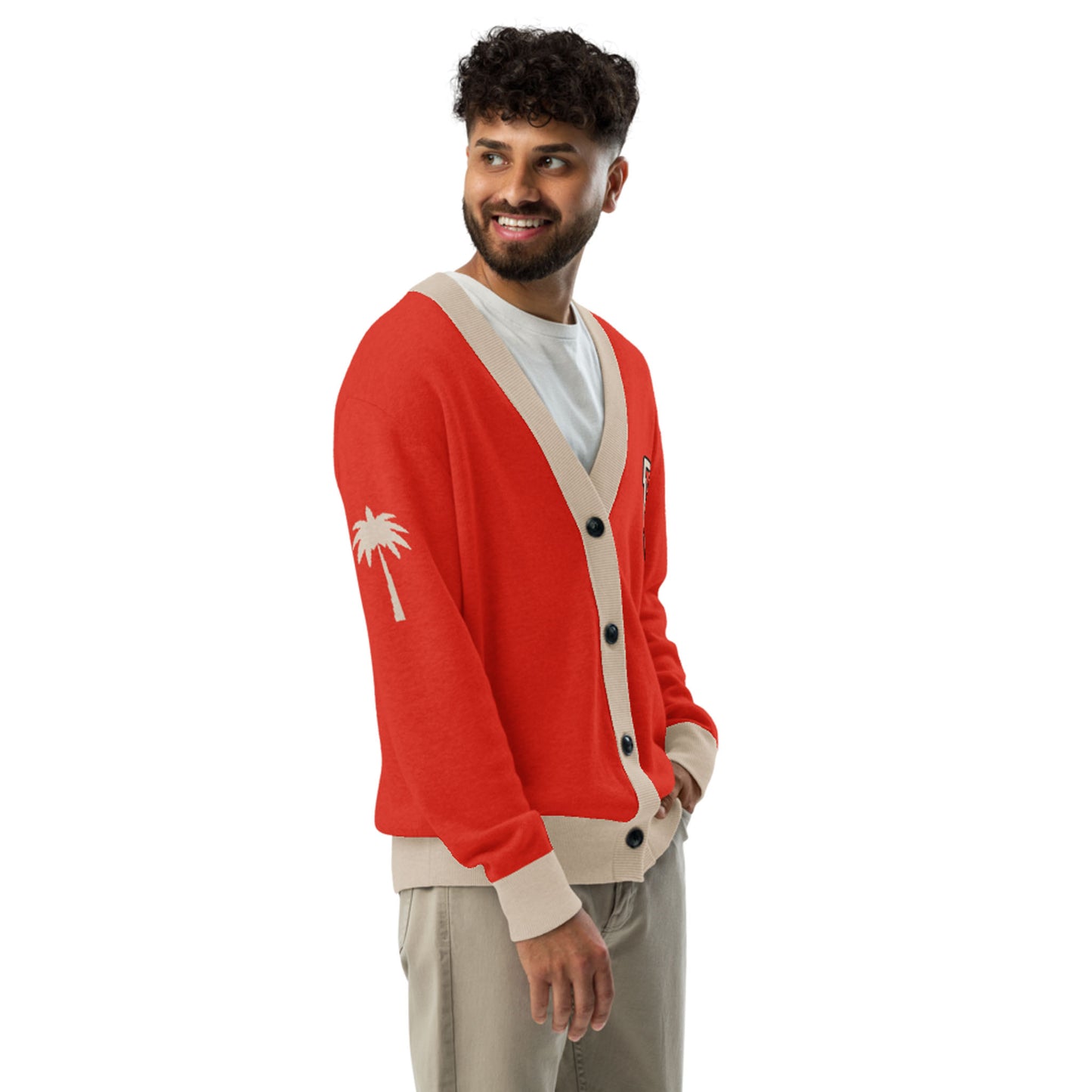 A smiling man with curly hair wears a two-tone cardigan with beige trim, a white shirt, and beige pants. The unisex cardigan features a white palm tree design on the left sleeve as he stands with his hands in his pockets, looking to the side.
