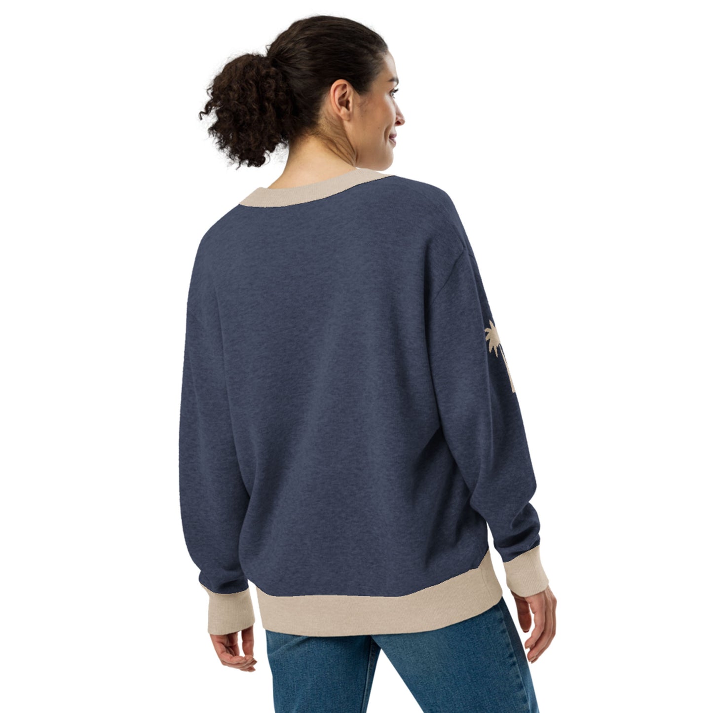 A woman with curly hair in a ponytail is wearing a loose, two-tone cardigan with beige cuffs and hem, paired with blue jeans. She is standing and facing away, looking over her shoulder.