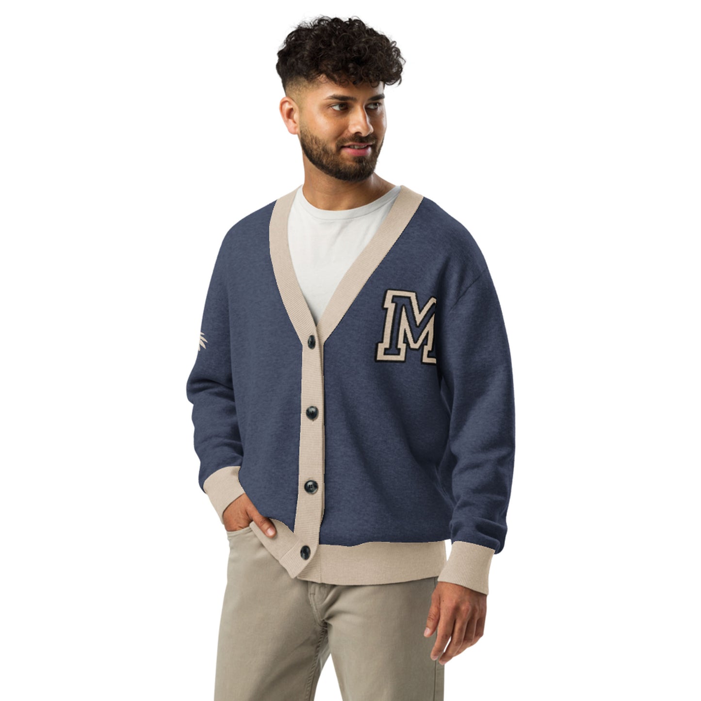 A man with curly hair and a beard wears a blue two-tone cardigan with beige trim, a large letter M patch on the chest, a white t-shirt, and beige pants, standing and looking to the side.