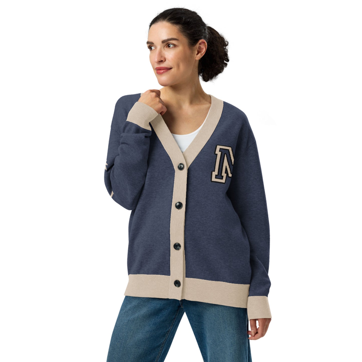 A woman in a two-tone jacquard knit cardigan with beige trim and an A patch stands with one hand on her collar, gazing to the side. Her dark hair is tied back, and she pairs the unisex cardigan with blue jeans and a white top.