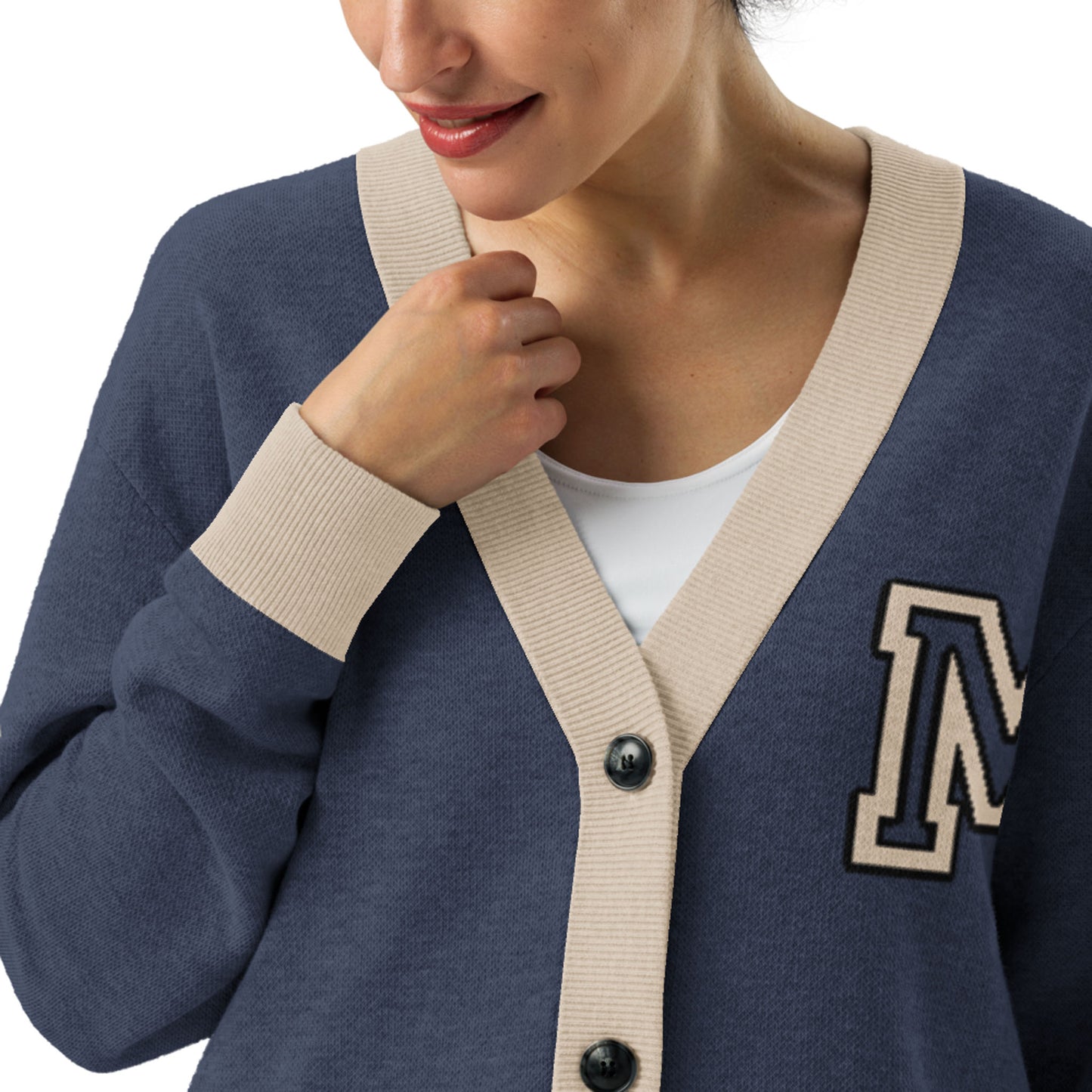 A woman wearing a navy blue jacquard knit cardigan with beige trim and an “M” patch on the chest over a white top, holding the edge of her cardigan and smiling.