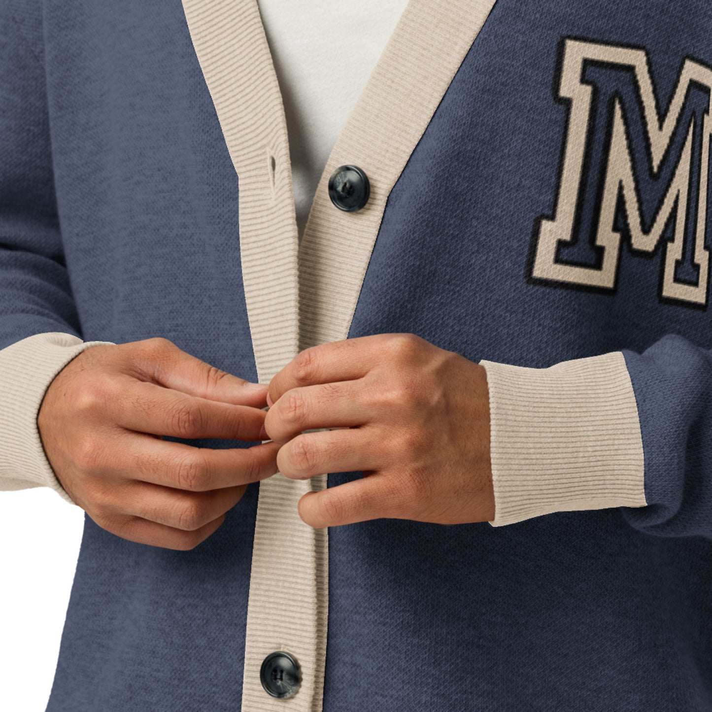 A person buttoning a soft jacquard knit cardigan in blue and beige with large black buttons and a white letter M patch on the chest. Only the hands and torso are visible.