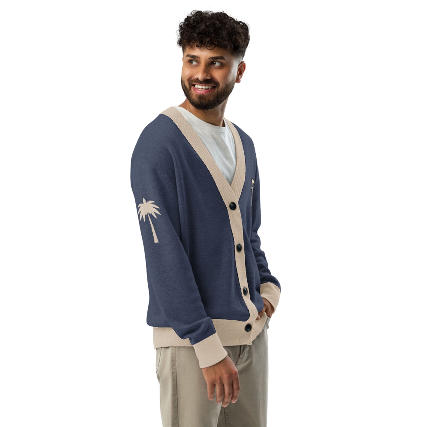 A man with curly hair and a beard smiles in a jacquard knit cardigan featuring beige trim and a palm tree design, layered over a white shirt and khaki pants, standing relaxed with his hands in his pockets.