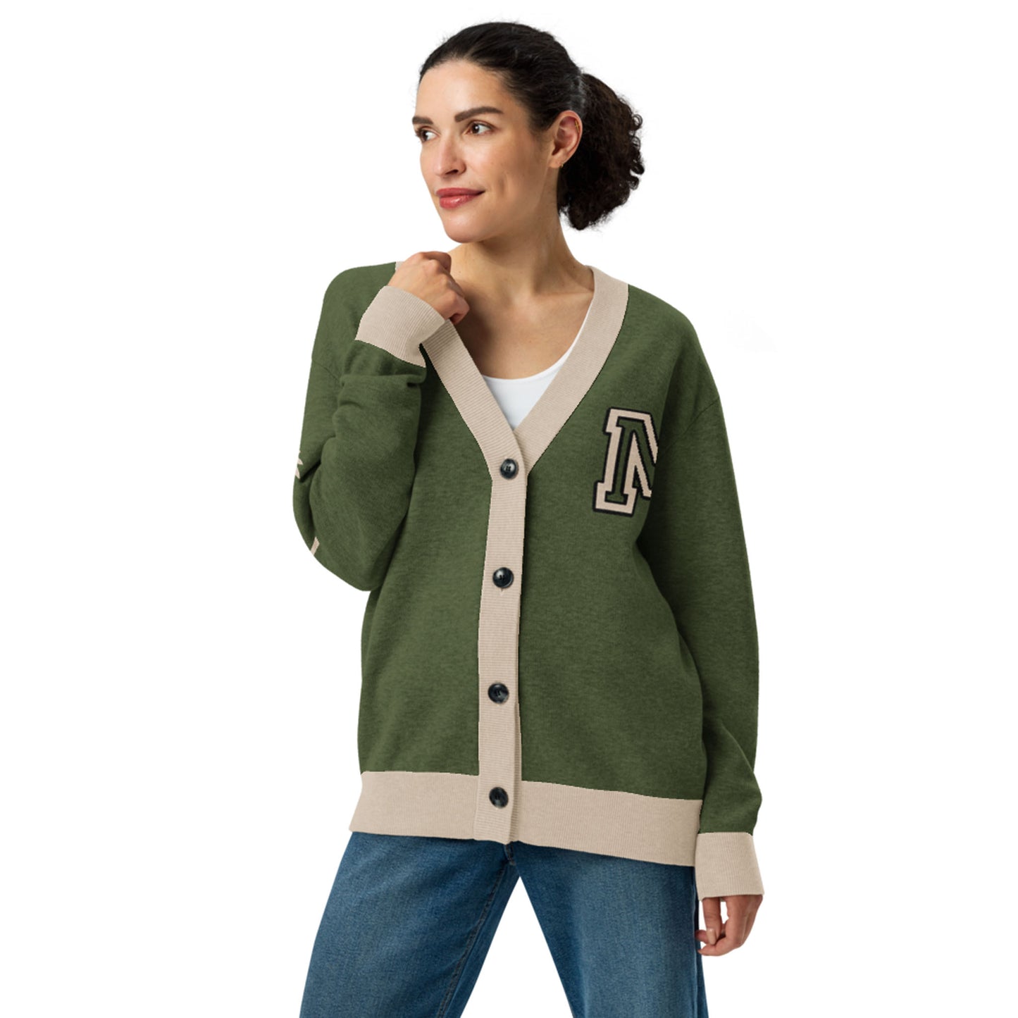 A woman wearing a two-tone cardigan with beige trim and buttons, featuring a large letter A patch on the left chest, over a white top and blue jeans, stands looking to the side with a slight smile.