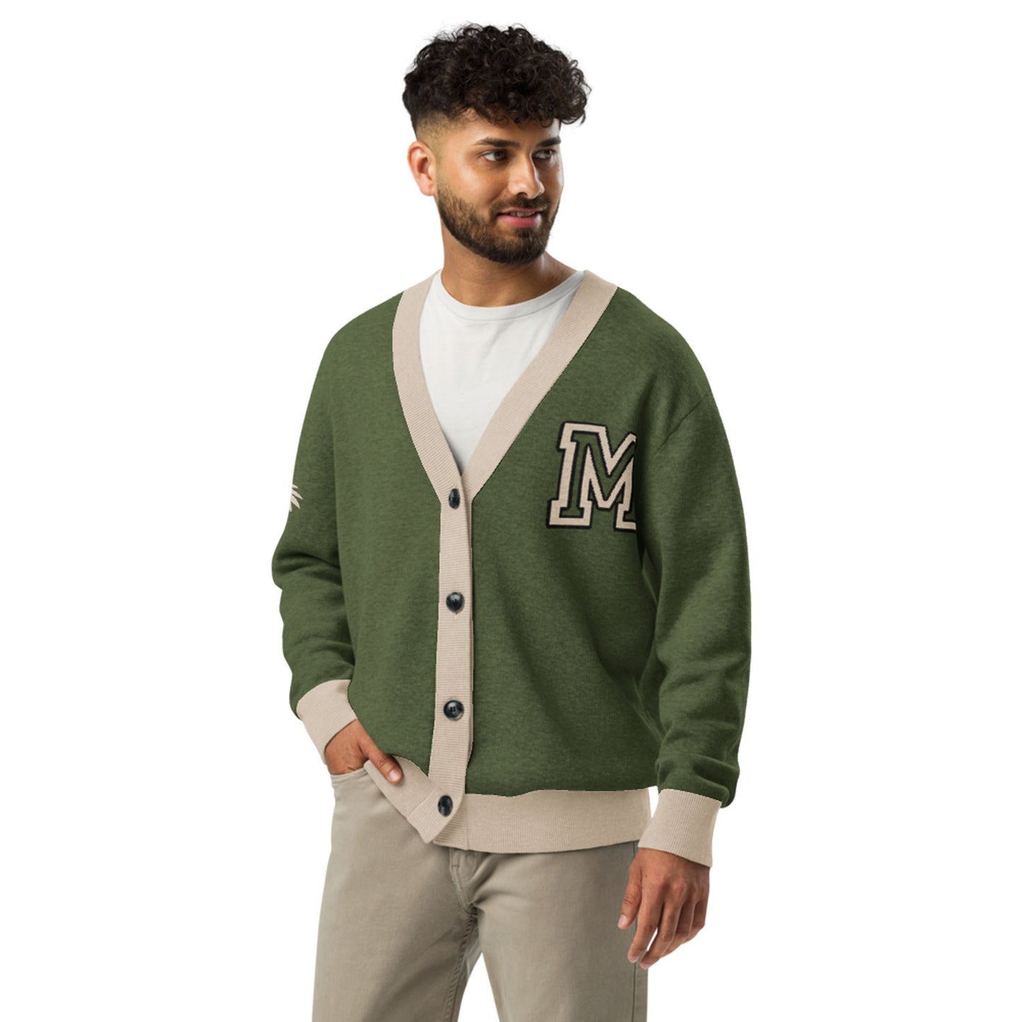 A man with curly hair and a beard wears a green jacquard knit cardigan with beige trim and a large letter M on the chest over a white t-shirt and beige pants, standing against a white background.