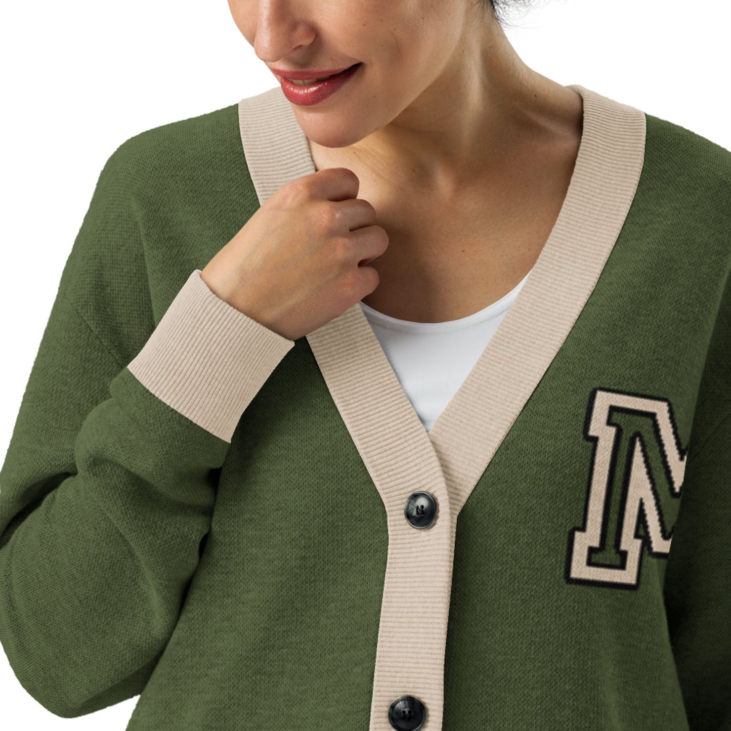 A woman wears a two-tone cardigan featuring a green jacquard knit with beige trim and an M patch on the chest, over a white top. She smiles slightly as she holds the edge of her cardigan near the collar.