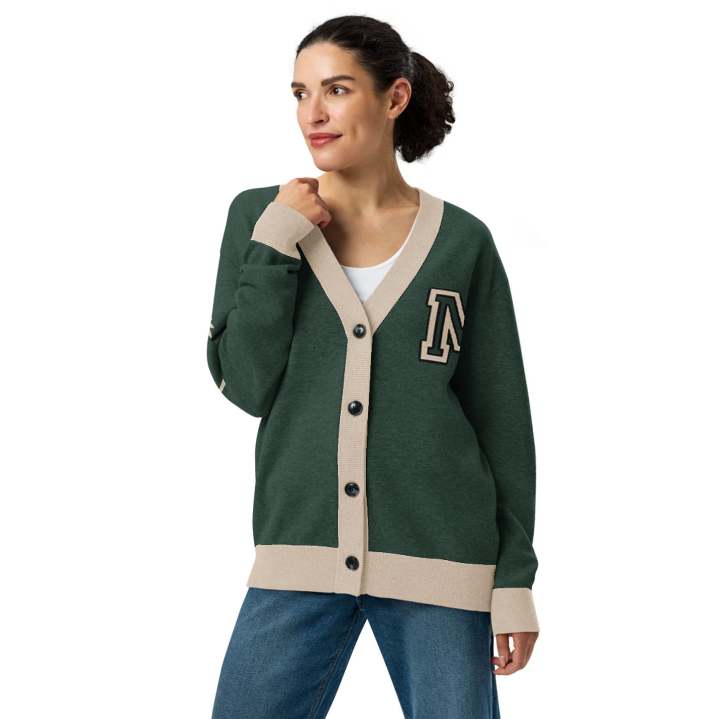 Woman wearing a green two-tone cardigan with beige trim and black buttons, featuring a large letter A patch on the left chest, white shirt underneath, and blue jeans, standing and looking to the side with a slight smile.