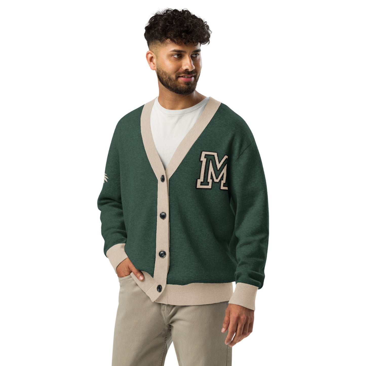 A man with curly hair and a beard wears a green and beige two-tone cardigan with a large M patch, a white T-shirt, and beige pants. He stands with one hand in his pocket, looking to the side and smiling.