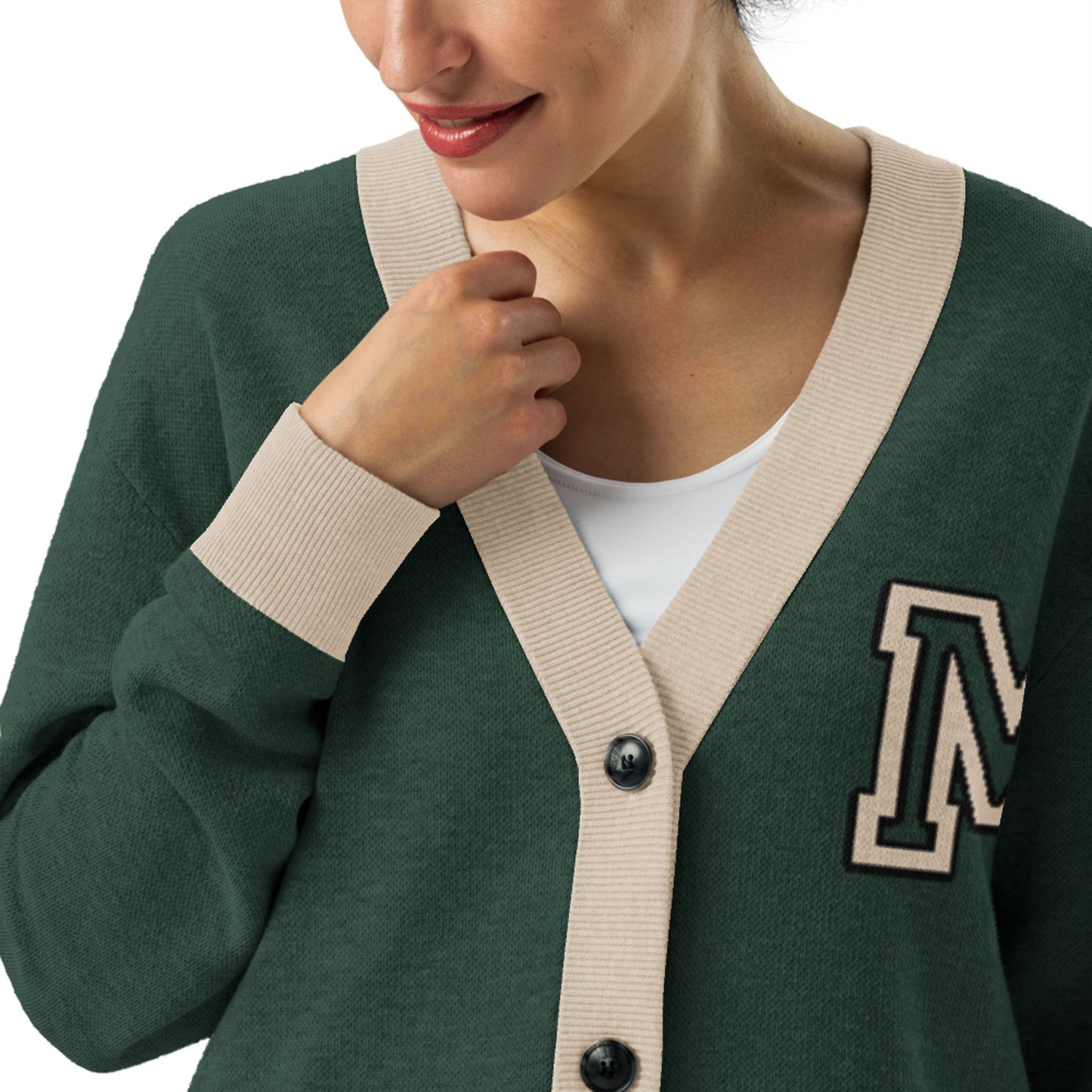 A woman wears a two-tone cardigan—a dark green style with beige trim and an M patch—over a white top, her hand gently holding the edge. Her face is partially visible, smiling softly.