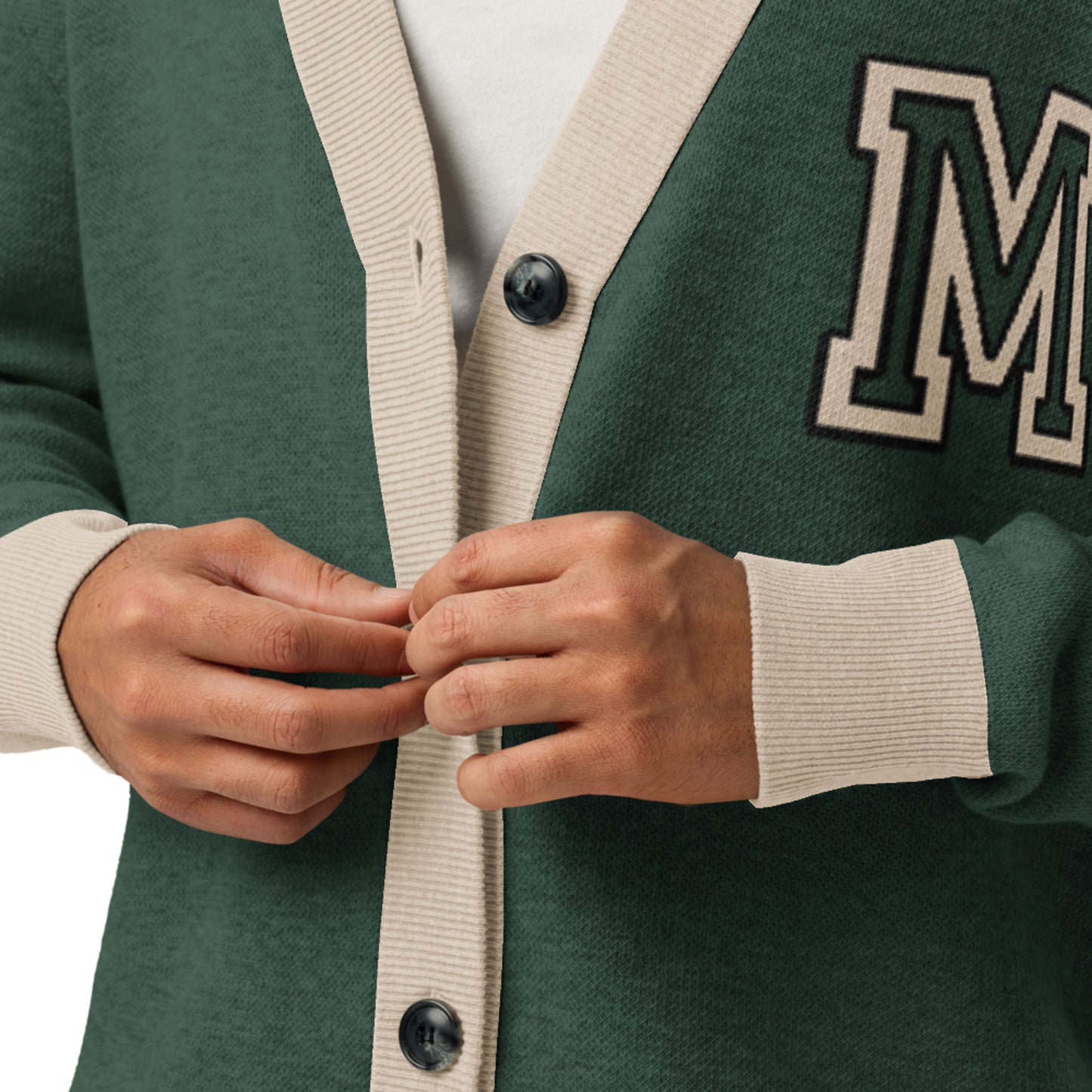 A person buttons a two-tone cardigan in dark green with light beige ribbed trim and large black buttons. This unisex cardigan features a bold letter M patch on the chest.