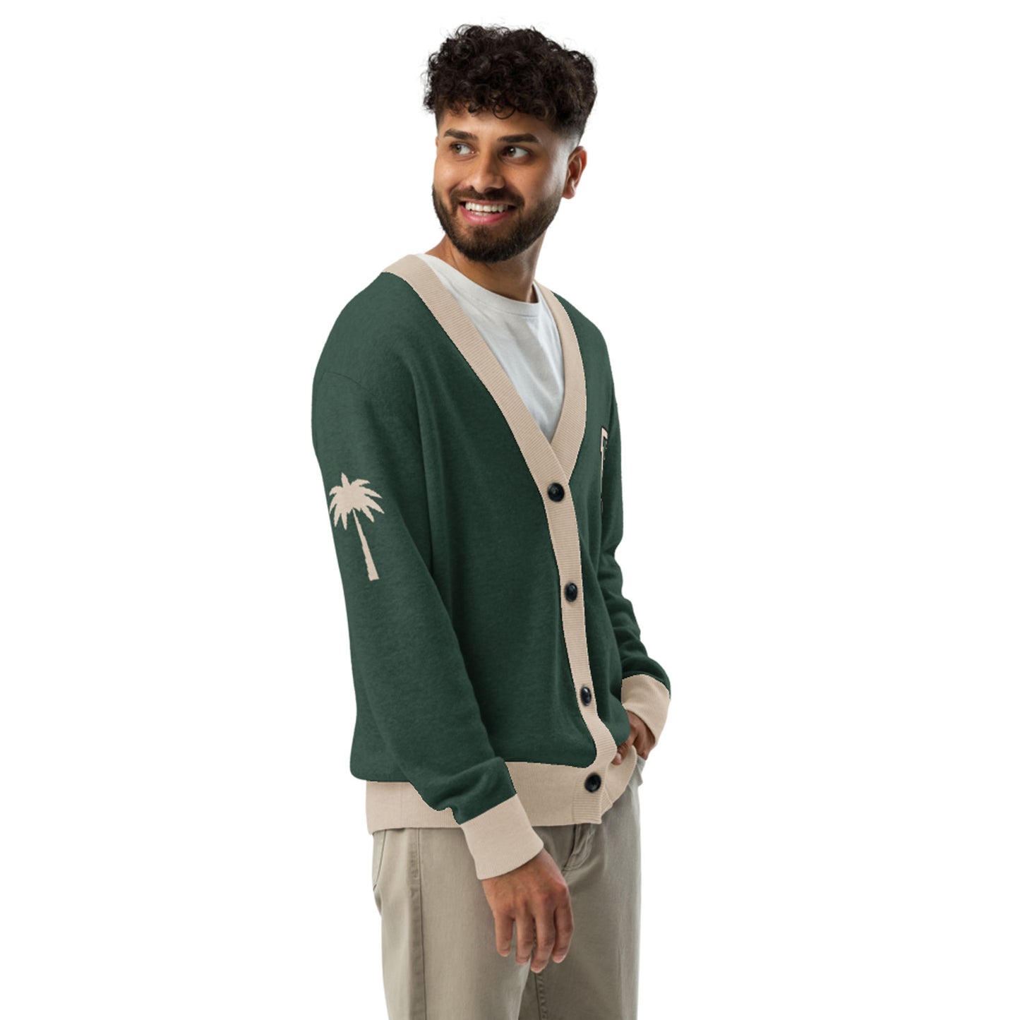 A man with curly hair and a beard wears a unisex two-tone cardigan featuring a palm tree design on the sleeve, paired with a white t-shirt and beige pants. He smiles warmly while looking to the side.
