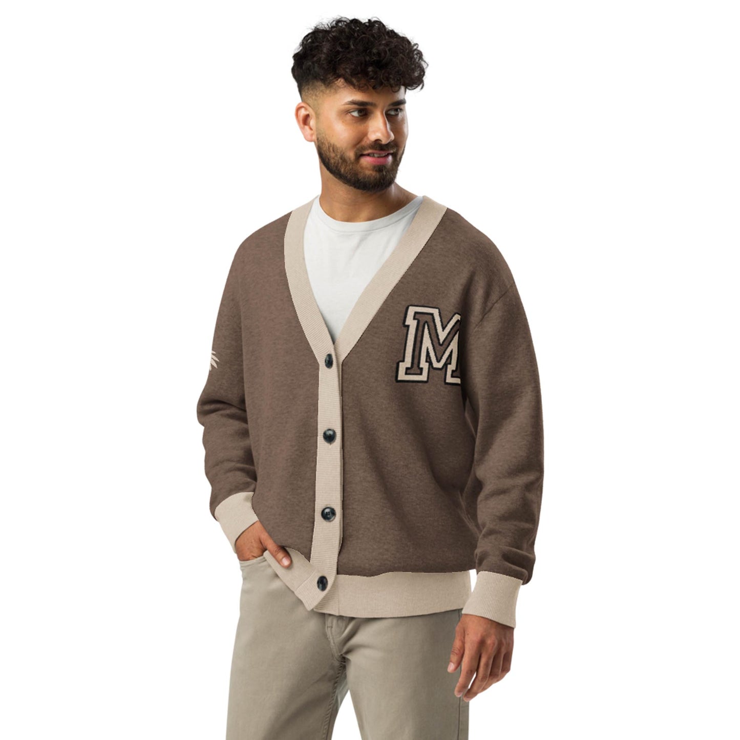 A man with short curly hair and a beard wears a two-tone jacquard knit cardigan with beige trim and a large letter M on the chest, over a white t-shirt and beige pants. He stands facing slightly right, smiling.