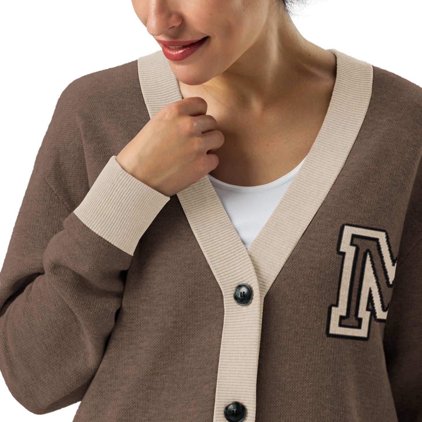 A woman wears a two-tone cardigan with beige trim and an M patch over a white top. She smiles slightly, gently holding the edge of her cardigan near her collarbone.