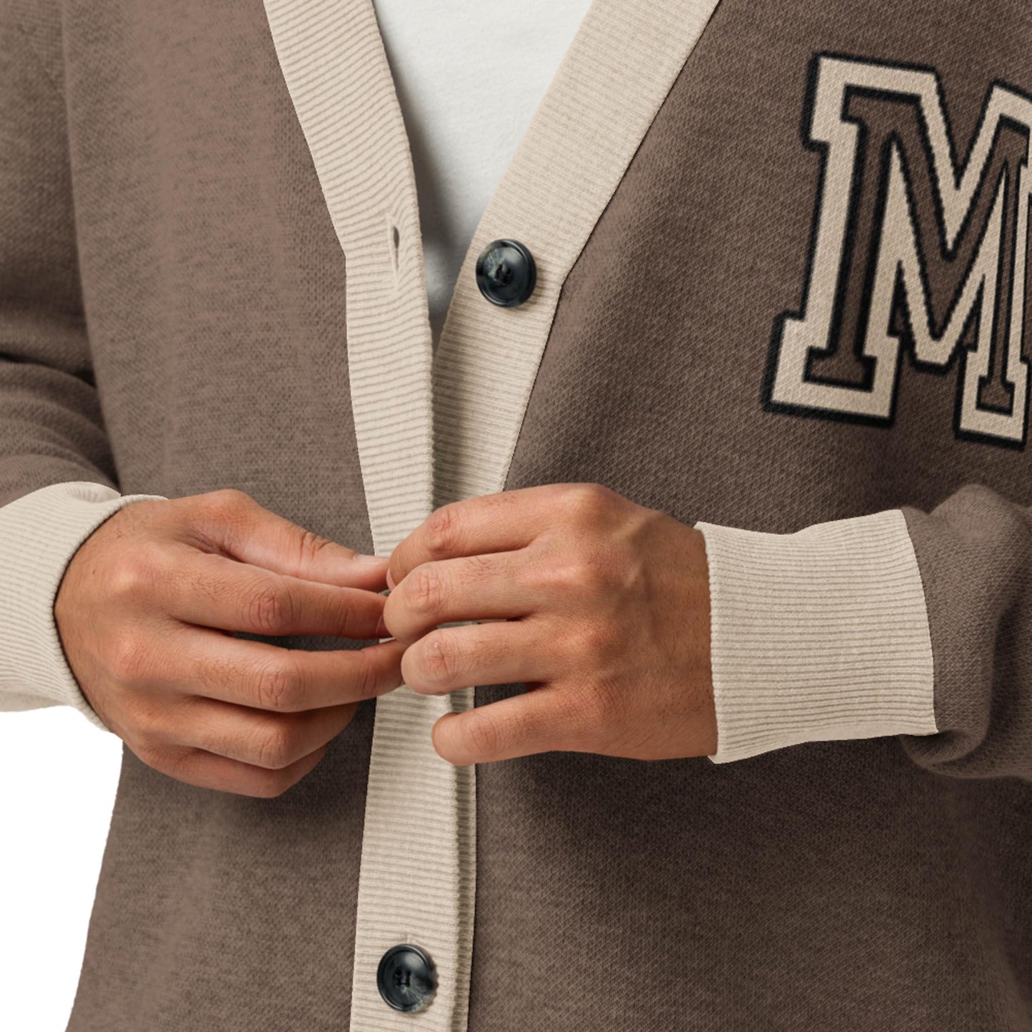 A close-up of a person buttoning a two-tone cardigan with large buttons and an embroidered letter M patch on the chest. This unisex cardigan, worn over a white shirt, features stylish brown and beige colors.