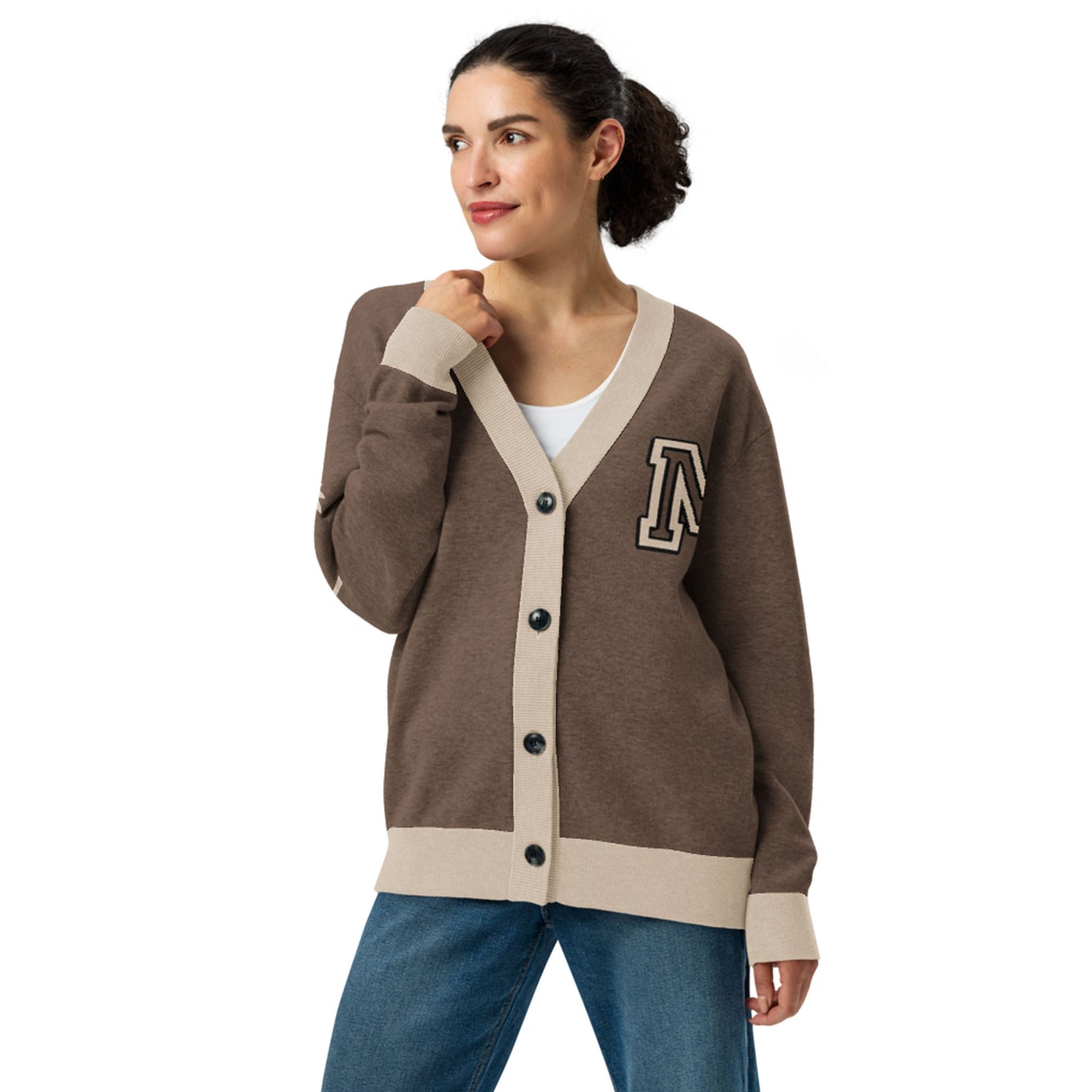 A woman wearing a two-tone cardigan with beige trim and black buttons, featuring a large letter A patch on the chest, stands with one hand on her collar and smiles while looking to the side.