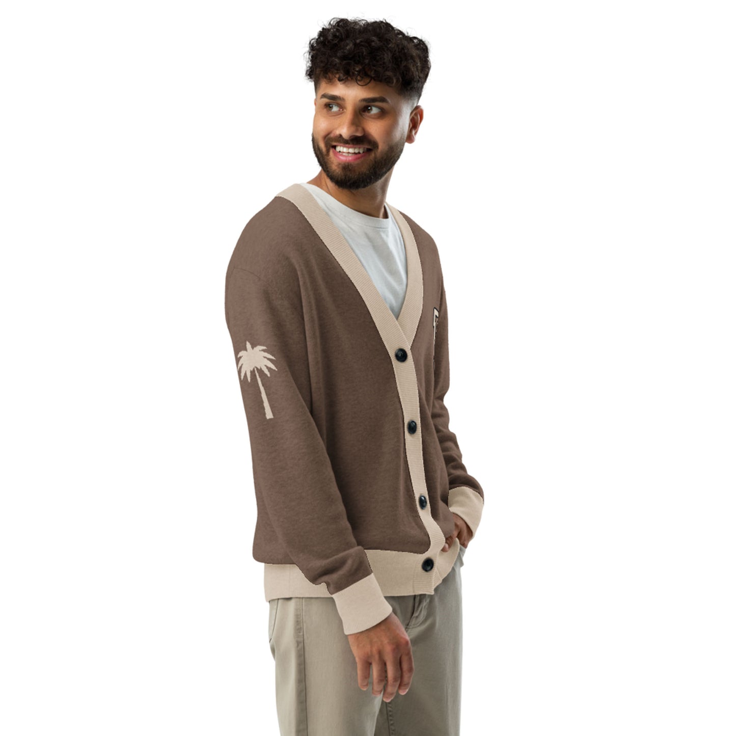 A man with curly hair and a beard, wearing a two-tone unisex cardigan with beige trim and a white palm tree design on the sleeve, stands sideways and smiles while looking to his left. He pairs it with a white shirt and light khaki pants.