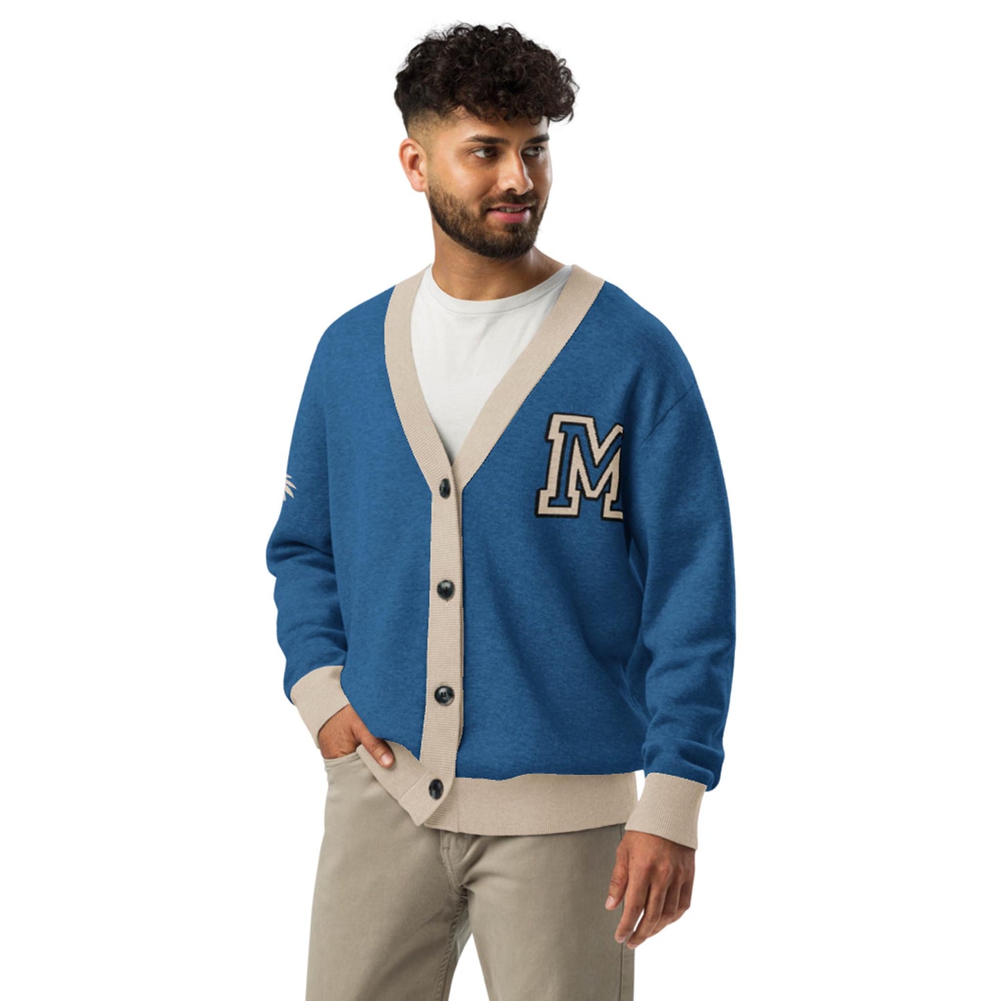 A man with curly hair and a beard wears a blue and beige two-tone cardigan with an “M” patch on the chest. The unisex cardigan is buttoned in front as he stands, one hand in his pocket, looking to the side and smiling.